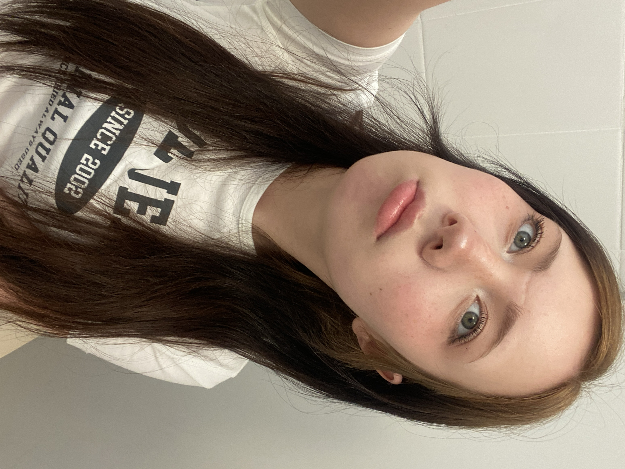 A selfie of a young woman with long brown hair and light eyes. She is wearing a white t-shirt with black lettering. The background is a plain white wall. The location is Unknown, Unknown.