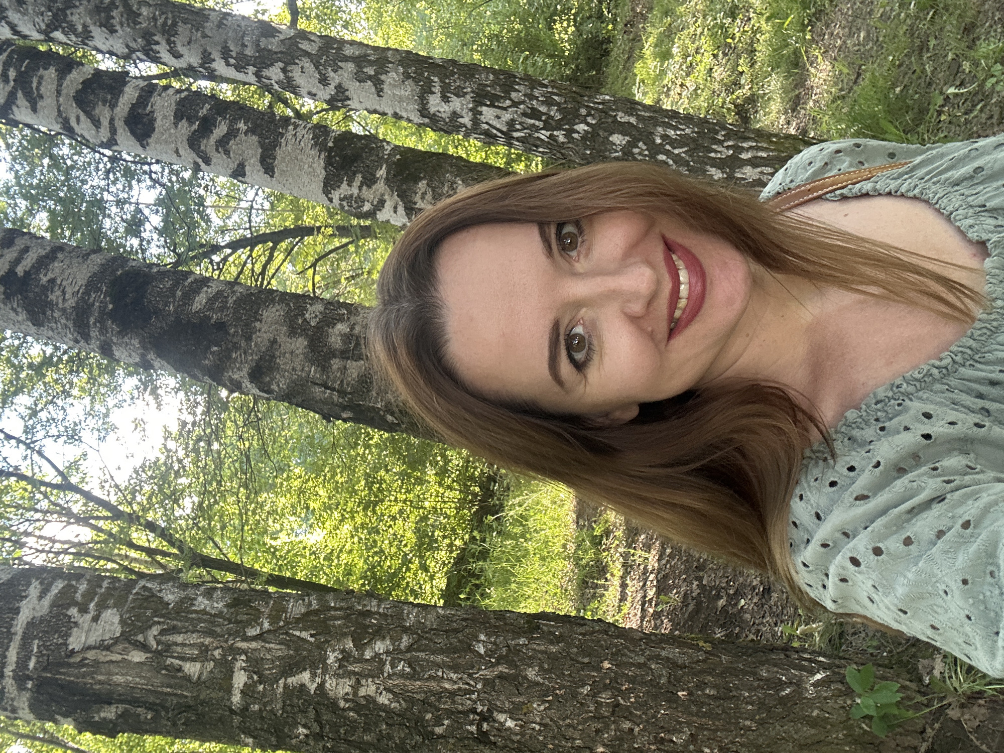 A woman with shoulder-length brown hair and light skin, wearing a light green, eyelet-embroidered blouse, is taking a selfie outdoors. She has on dark red lipstick and is smiling broadly, revealing her teeth. The background consists of birch trees with their characteristic white and black bark, interspersed with lush green foliage and sunlight filtering through the leaves. The ground is covered with dry leaves and grass. The scene appears to be a park or forest area. The location is stated to be in Medvedevo, Russia.