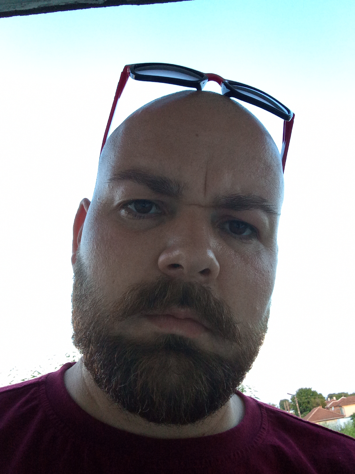 A close-up, upward-angled photograph shows a bald, fair-skinned man with a full beard and mustache. He has sunglasses perched on top of his head. His facial expression appears neutral, possibly contemplative. He is wearing a maroon or burgundy-colored crew neck shirt. The background shows a bright, possibly overcast sky. In the lower right corner of the image, rooftops of buildings and trees are faintly visible, suggesting an outdoor setting. Based on the prompt, the location is Roza, Bulgaria, although specific details to confirm the city are absent from the image itself.