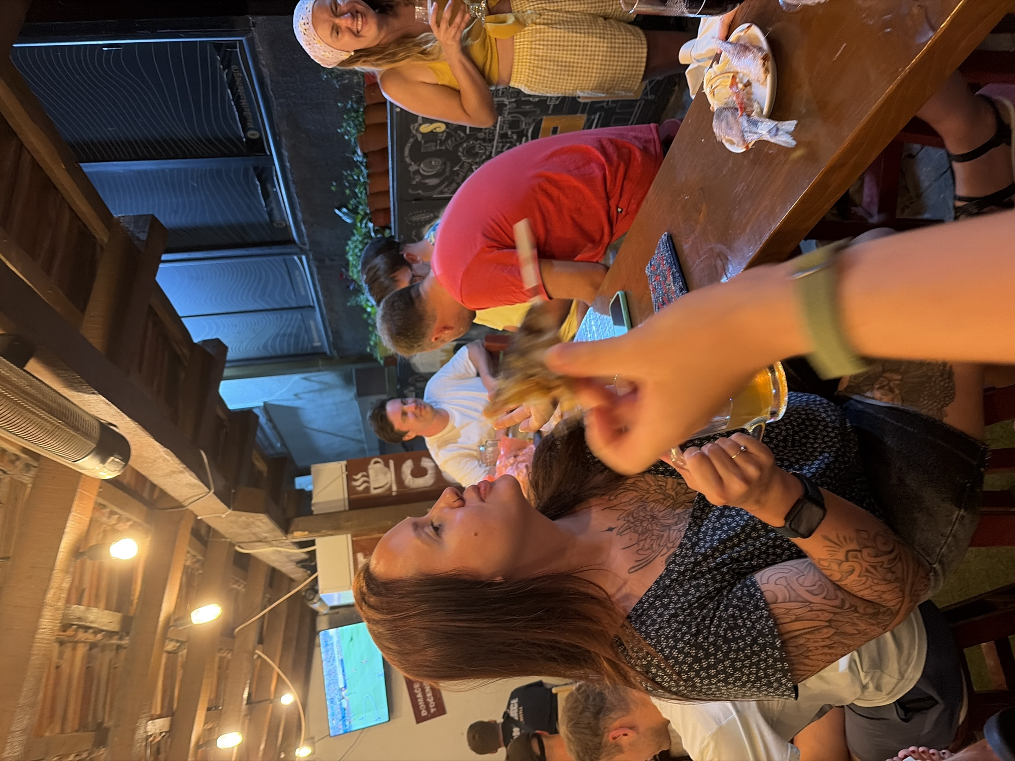 A group of people are gathered around a wooden table at an outdoor establishment in Bar, Montenegro. The scene is illuminated by overhead lighting and a television screen displaying a soccer match. In the foreground, a woman with long brown hair and extensive tattoos on her arms and chest is holding a piece of food and a glass of beer. To her right, a woman in a yellow top and checkered shorts laughs and holds a glass. Other individuals are visible at the table, some eating and drinking. The environment suggests a casual dining or bar setting, with plants visible in the background and exposed wooden beams overhead.