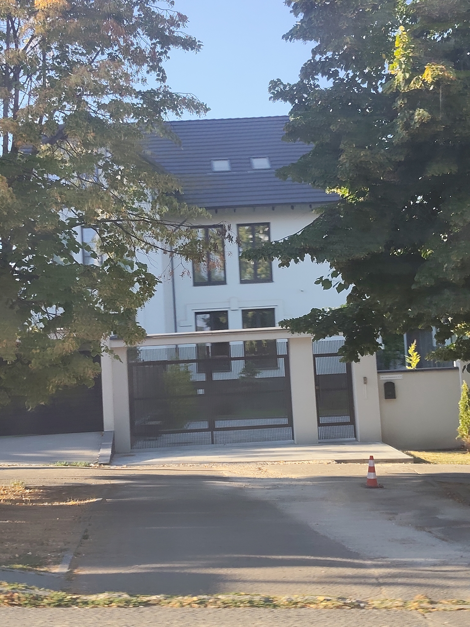 The image shows a modern white house with a dark gray pitched roof and several large windows, some of which reflect the surrounding trees and sky. A black metal gate with vertical bars and a section of corrugated metal is visible in front of the house. The gate is flanked by two white pillars, and there is a paved driveway leading up to it. A single orange and white traffic cone stands on the driveway. The house is partially obscured by the branches of trees with green and yellowing leaves, suggesting it is autumn. The sky is clear and blue. The scene appears to be a residential property.