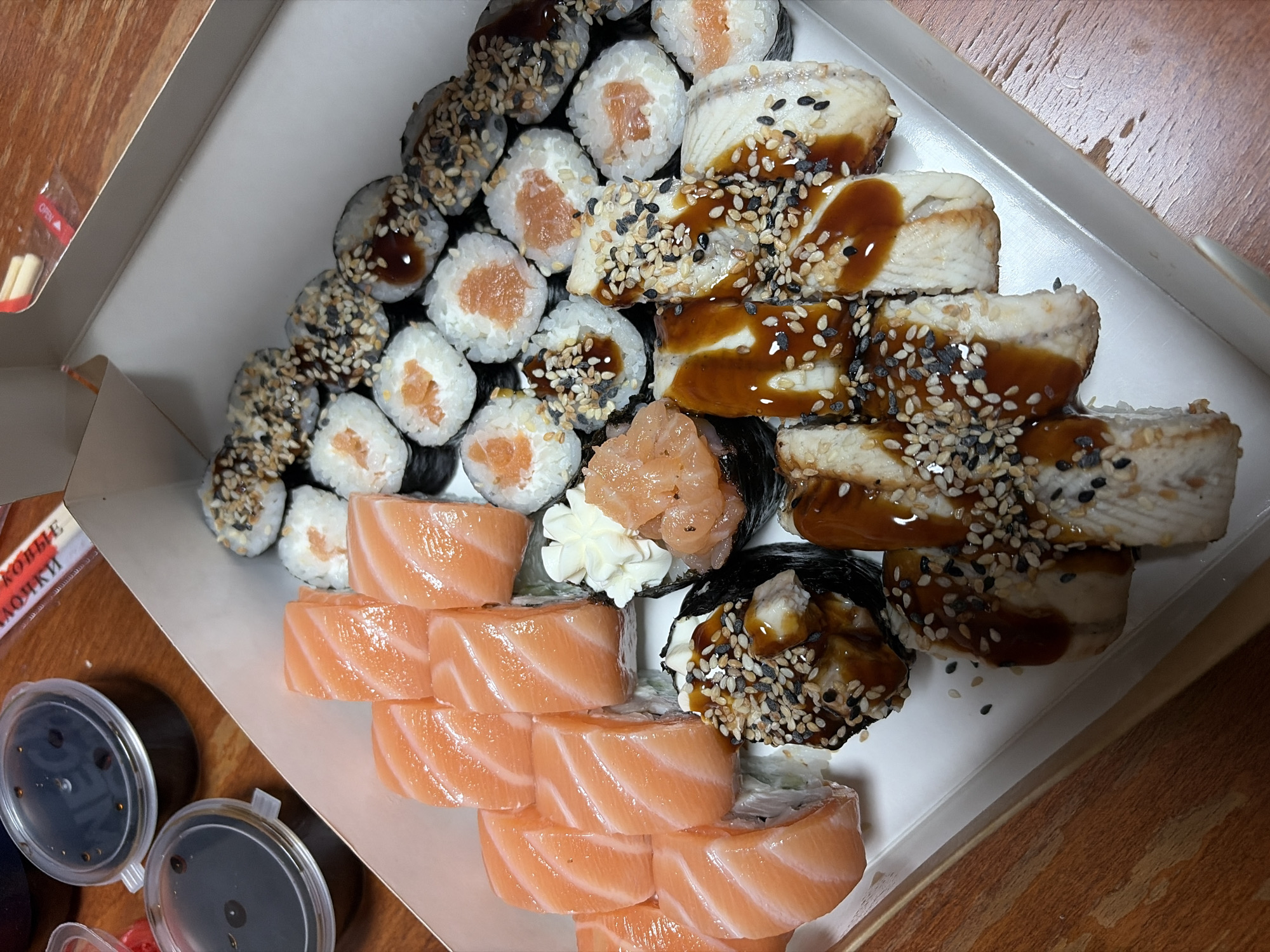 The image shows a white cardboard takeout box filled with a variety of sushi and sashimi. The sushi includes pieces with raw salmon on top, some with sesame seeds and a dark sauce, and others with eel and a similar sauce. There are also some smaller maki rolls with visible salmon. A white dollop of what appears to be wasabi or cream cheese is visible on one of the sushi pieces, and a small mound of what looks like minced salmon with seasoning is also present. The box is placed on a wooden surface, likely a table. In the upper left corner, two small, clear plastic containers with dark lids, likely containing soy sauce, are visible. A portion of red and white chopsticks in a clear plastic wrapper is also present. The scene is shot from a high angle looking down into the box. The context of Chaikovsky, Russia, is not directly evident from the visual information within the image itself.