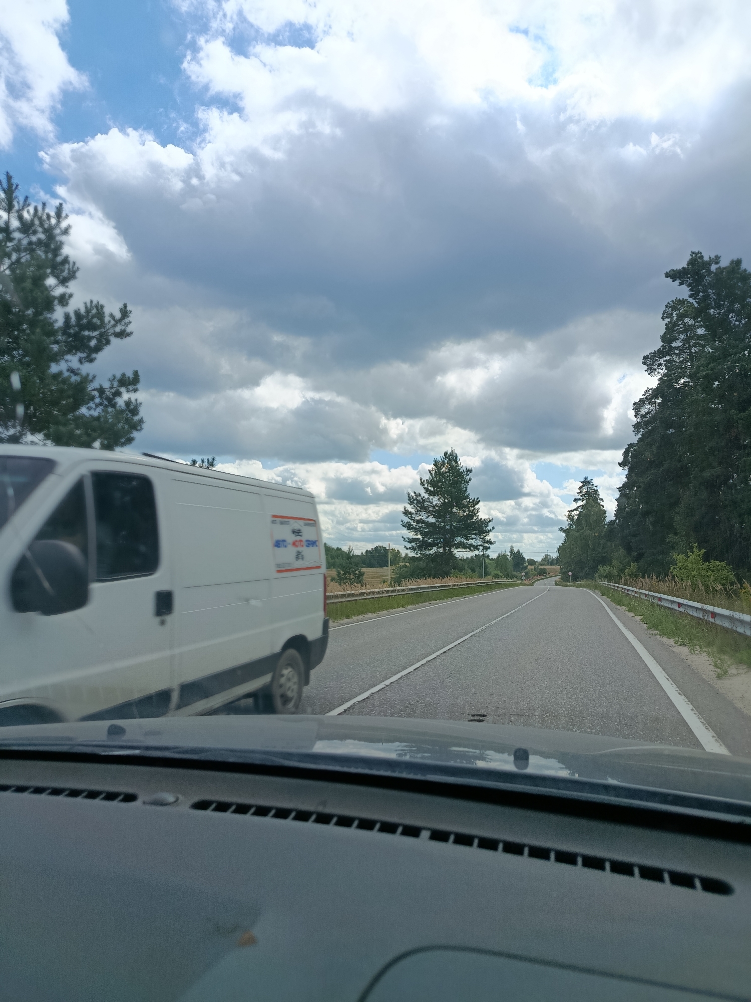 A white van is seen driving on a two-lane road in Gribchikha, Russia. The van is on the left side of the road. The road is paved and has white lines marking the lanes. The scene is under a partly cloudy sky. On the right side of the road, there is a guardrail and trees. The van has some text on its side, but it is difficult to read. The image is taken from inside a vehicle, looking out the windshield. The dashboard of the vehicle is visible in the foreground.