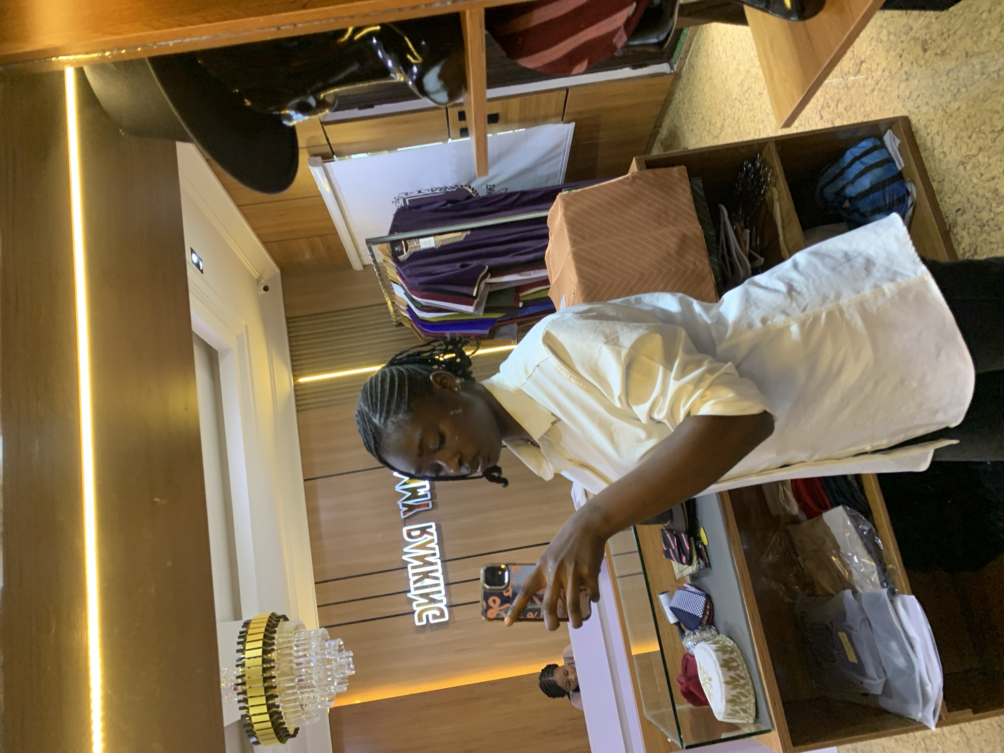 The image depicts the interior of a retail establishment, possibly a clothing store, in Gura Top, Nigeria. In the foreground, a young woman with braided hair is holding a smartphone and appears to be taking a selfie or recording a video. She is wearing a white collared shirt and dark pants. To her left, a shelf displays various accessories, including a white cap with gold embroidery, ties, and patterned handkerchiefs. Below this shelf, folded garments are visible in plastic packaging.
In the background, a rack of clothing, including several dark purple garments with intricate gold patterns, is displayed. A mannequin wearing a dark hat is partially visible to the right of the frame. The store features wooden paneling on the walls, a chandelier, and neon lighting. Another person is visible in the reflection of a mirror or glass surface in the background, behind the counter. The overall impression is a well-lit retail space focused on fashion or traditional attire.