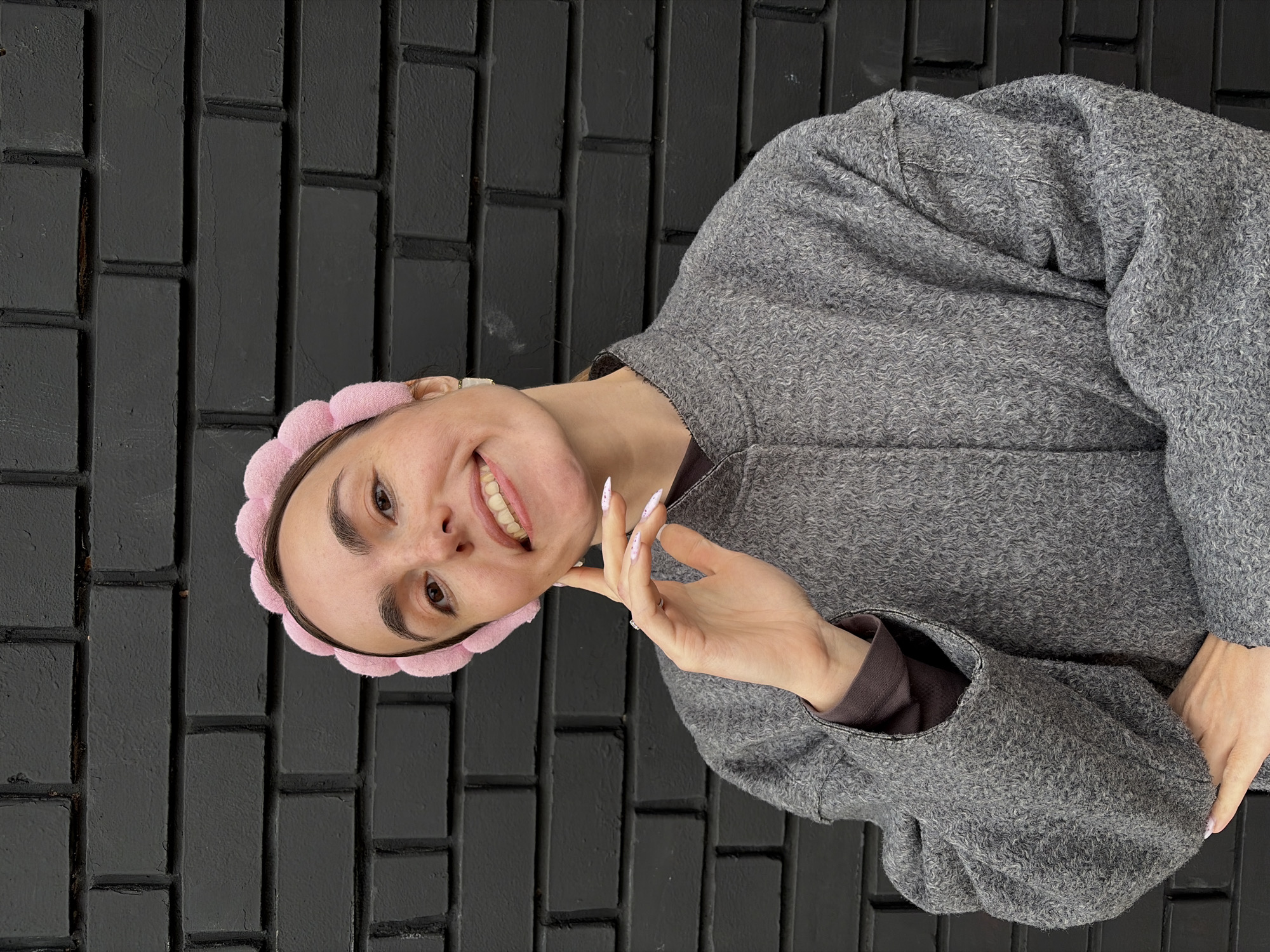 A young woman is positioned at an angle in front of a black brick wall. She wears a gray, textured jacket, a pink headband, and has long, white manicured fingernails. She is smiling and touching her chin with one hand. The background appears to be an indoor brick wall. There is no obvious evidence in the image to confirm that this is in Izhevsk, Russia.