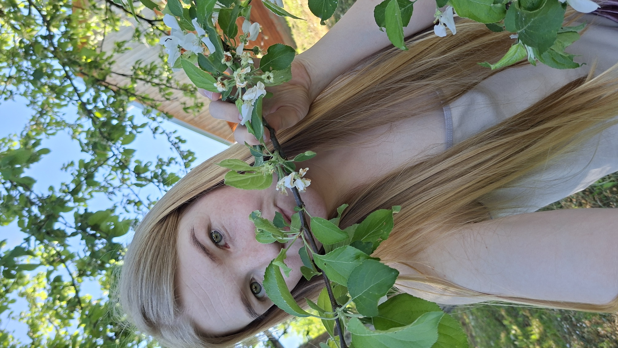 The image shows a woman with long blonde hair, holding a branch with white flowers and green leaves. The background features a clear blue sky with trees and a building visible. The photograph appears to be taken outdoors, possibly in a garden or park. The location cannot be confirmed without additional information.