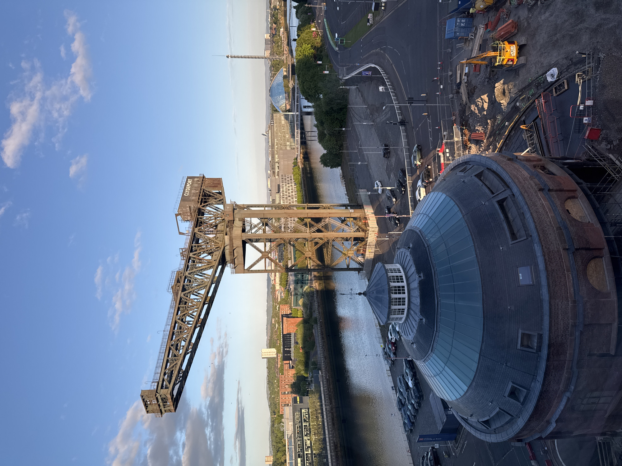 This photograph, taken from an elevated position, showcases a scene in Glasgow, United Kingdom. The image is dominated by the Finnieston Crane, a prominent steel cantilever crane, set against a blue sky with scattered clouds. The River Clyde runs beneath the crane, reflecting the surrounding cityscape and sky.
To the right of the crane is a large, circular brick building with a dome-shaped glass roof. Cars are parked near the base of this building. The scene includes city infrastructure such as roadways, buildings, and a construction site with heavy machinery. The Glasgow Tower is visible in the background.