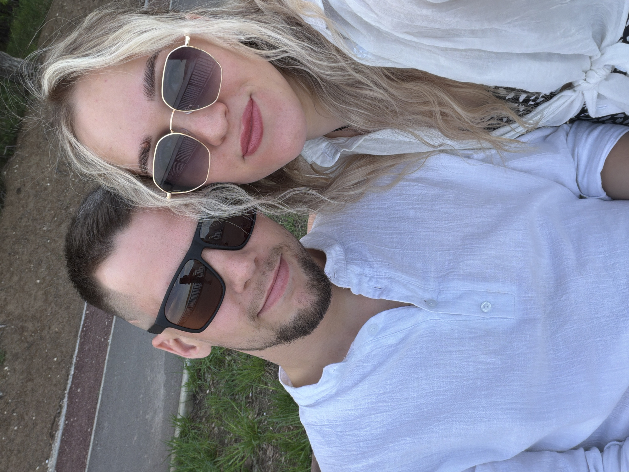 A selfie shows a woman and a man close together, likely in an outdoor setting. The woman, positioned towards the top of the frame, has blonde hair and is wearing sunglasses and a white shirt. The man, located at the bottom, has short dark hair, a beard, and is also wearing sunglasses and a white shirt. They are on a sidewalk in Unknown, Unknown.