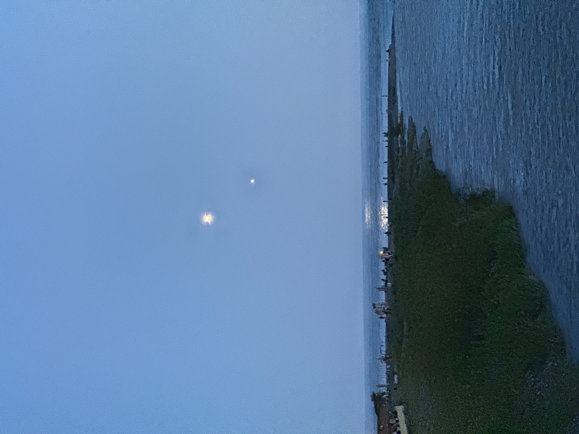 The image depicts a serene twilight or night landscape in Sochi, Russia, overlooking a vast body of water, likely the Black Sea. The scene is characterized by a deep blue sky that occupies a significant portion of the frame, indicating the late hours of the day.
Two prominent points of light are visible in the sky: a larger, brighter object, possibly the moon or a very luminous planet, which exhibits a distinct starburst effect, and a smaller, fainter light source positioned further away.
On the right, the dark expanse of the water shows subtle textures and reflections of artificial lights from the distant shore. The left side features a strip of land, possibly a coastline or embankment, covered in dark green, low-lying vegetation. Along the edge where the land meets the water, numerous small, bright artificial lights are illuminated, suggesting the presence of human infrastructure such as buildings, streetlights, or a promenade. These lights create distinct glimmers and reflections on the water's surface.
No specific activities are clearly taking place, and no individuals are distinctly visible. The weather appears clear with no clouds. The overall ambiance is peaceful and calm. There is no visible text in the image.