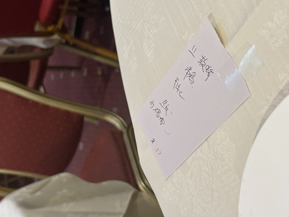 A close-up shows a light pink note taped to a tablecloth. The note is written in what appears to be Chinese characters. The characters are stacked in vertical columns, with some symbols underneath. The date "2.17" is written in red ink at the bottom of the note. The tablecloth is light beige with a subtle patterned texture. Red chairs with gold trim are visible in the background.