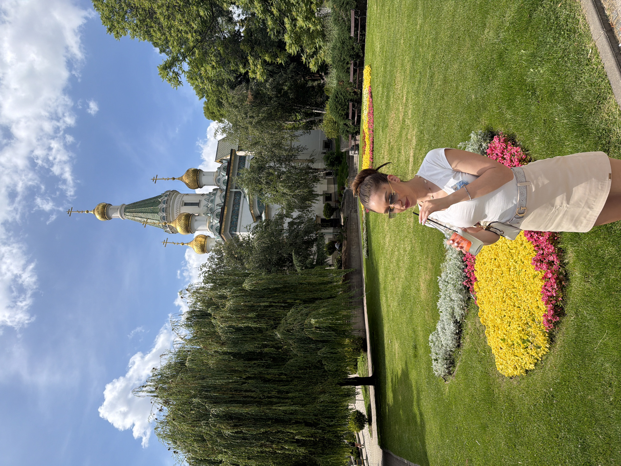 The image depicts a young woman standing in a park in Sofia, Bulgaria, with a distinct church in the background. The woman, wearing a white t-shirt, a beige skirt, sunglasses, and a belt with a prominent 'G' buckle, is holding a drink with a straw and appears to be taking a sip. Her hair is pulled back in a bun.
The park features a meticulously maintained lawn with colorful flowerbeds in shades of yellow, pink, and silver. Behind the flowerbeds, several dark brown park benches are visible. A large weeping willow tree stands to the left of the frame, its branches cascading down.
Dominating the background is the Saint Sofia Church, an iconic landmark in Sofia. The church is characterized by its white facade, green-tiled roof, and multiple golden domes topped with crosses. The architectural style suggests an Orthodox Christian church. The sky above is blue with scattered white clouds. The overall scene is bright and sunny, suggesting a pleasant day.