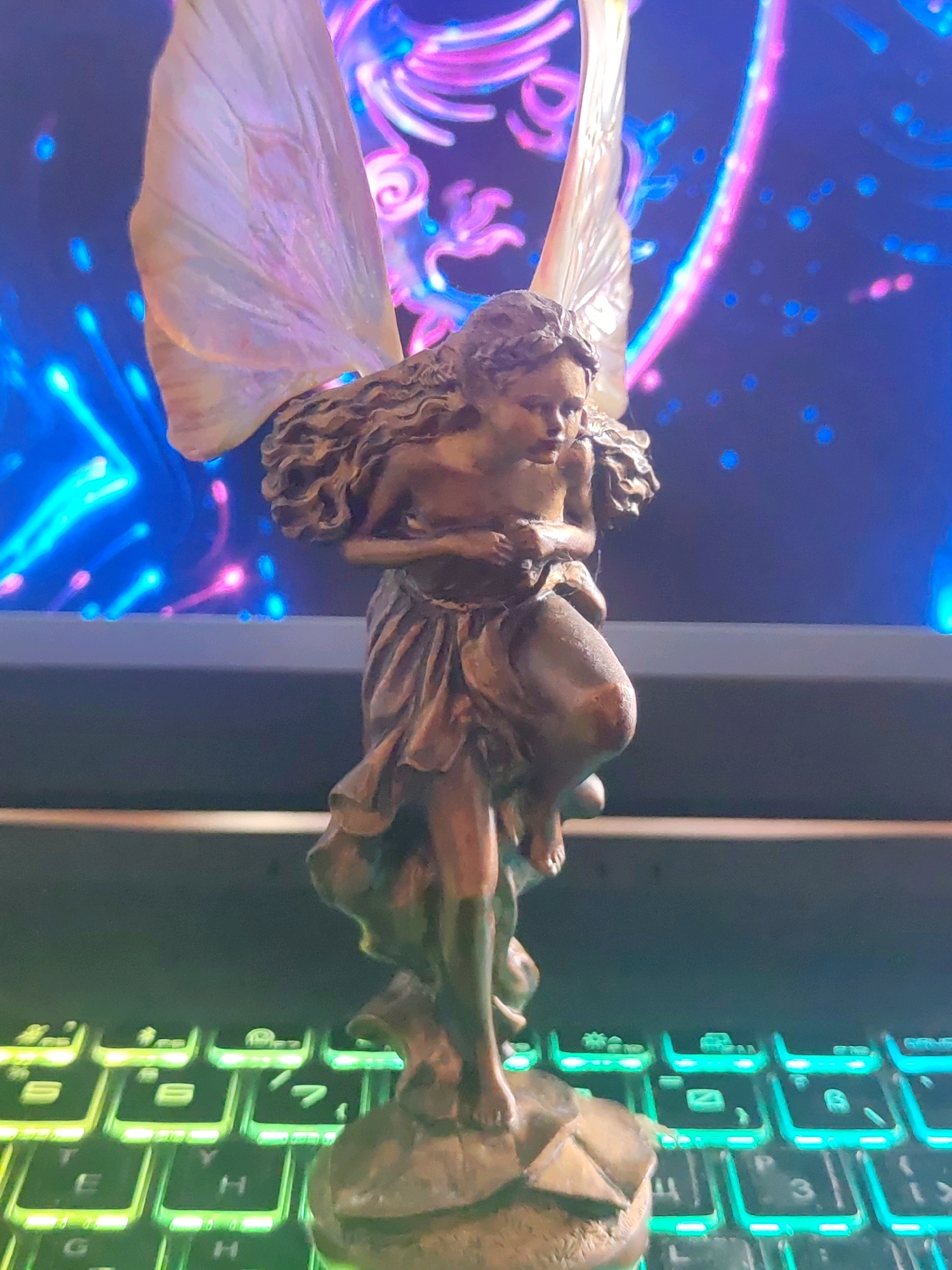 A bronze-colored statue of a winged figure, possibly a fairy or angel, stands on a pedestal in the foreground. The figure has long, wavy hair, a draped garment, and delicate wings that are translucent and have iridescent hues. The statue is positioned in front of a computer monitor displaying a vibrant, abstract image with swirling patterns of pink, blue, and purple light. Below the monitor, a backlit keyboard with green lighting is visible. The overall scene suggests a personal display or workspace. This image was captured in Schwerin, Germany.