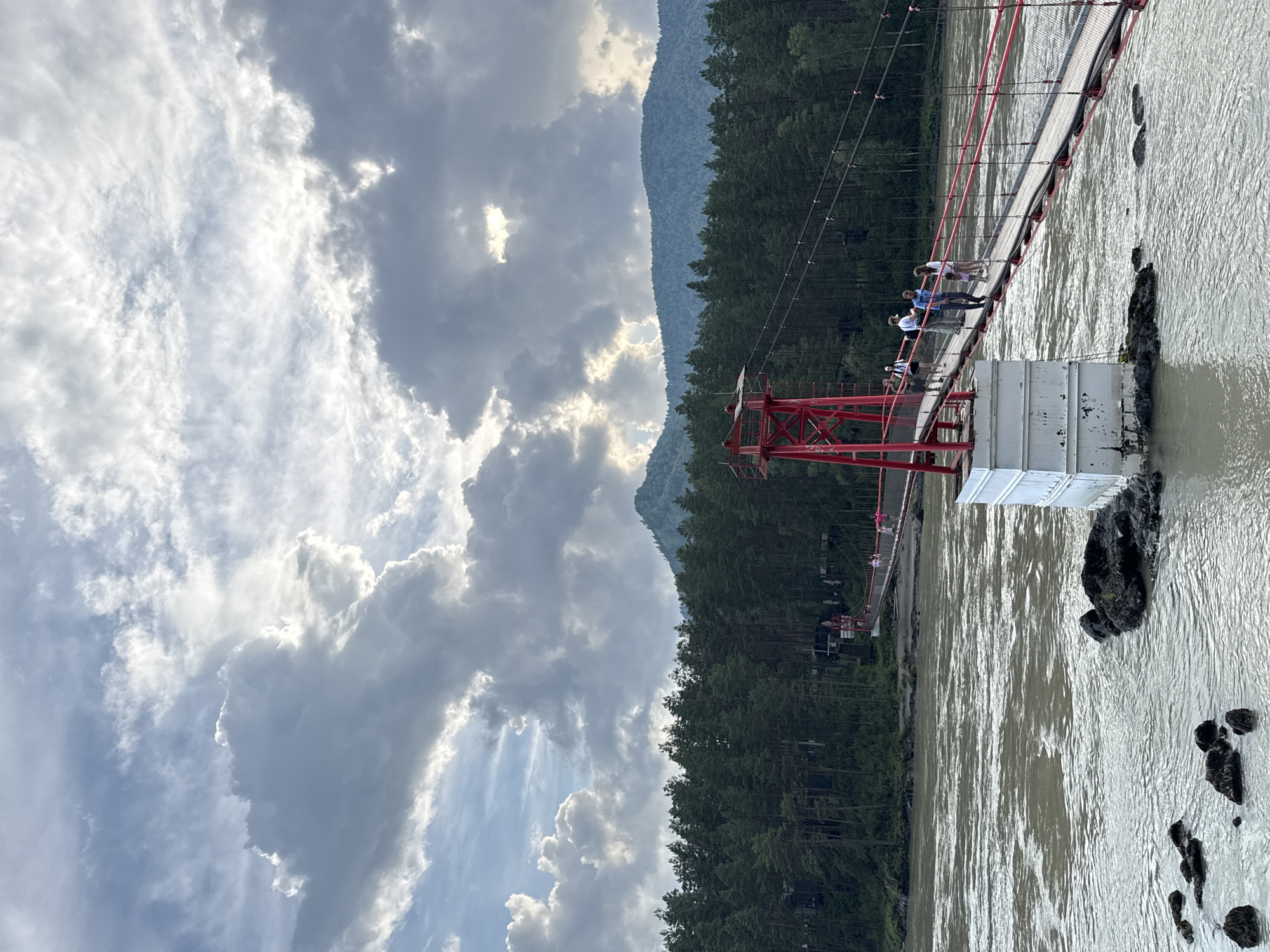 A suspension bridge, painted red and white, spans a wide, murky river. The bridge is populated by several people, some walking, others standing. A dense forest of evergreen trees lines the opposite bank, with mountains visible in the distance. The sky is filled with dramatic, grey clouds, with patches of sunlight breaking through. The location is the tourist area of the Katun River in the Altai Republic, near the village of Elekmonar and not far from the city of Gorno-Altaysk, Russia, though it is not located in the city of Barangol.