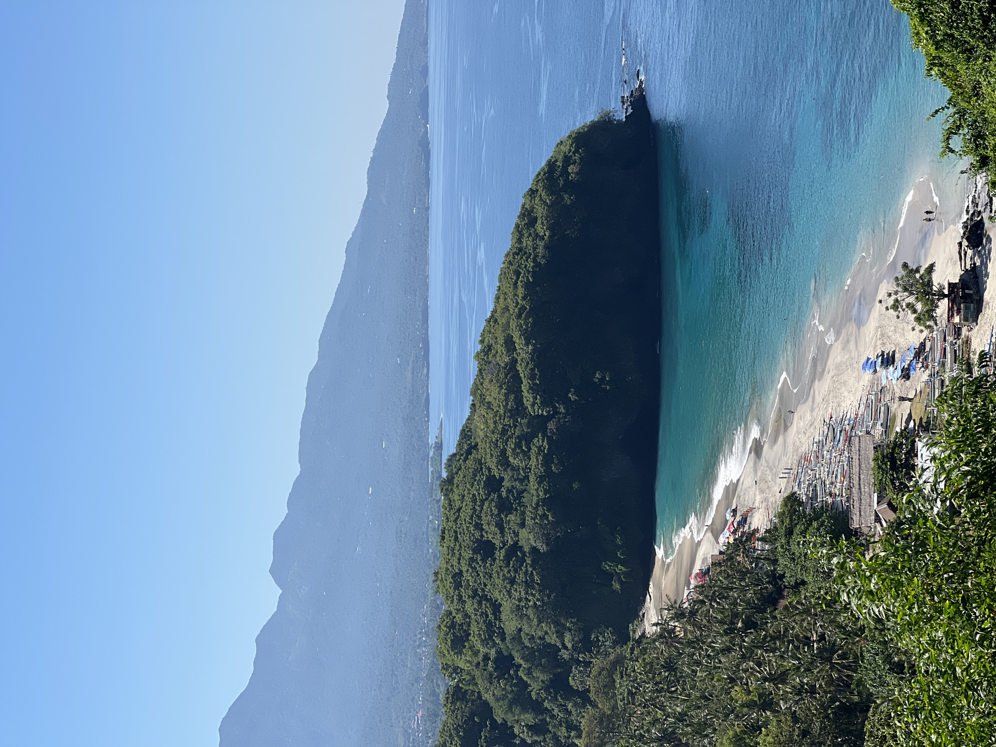 This is an aerial shot of Virgin Beach in Karangasem, Indonesia. The image displays a crescent-shaped sandy beach bordering a bright turquoise ocean. Numerous outrigger boats are lined up on the shore, some with colorful canopies. A few people are visible on the beach, some walking along the water's edge, and others resting under umbrellas. Lush green vegetation, including palm trees, covers the land leading up to the beach and extends to a large, tree-covered headland jutting into the sea. In the background, a range of hazy blue mountains forms the horizon, with a clear blue sky overhead.