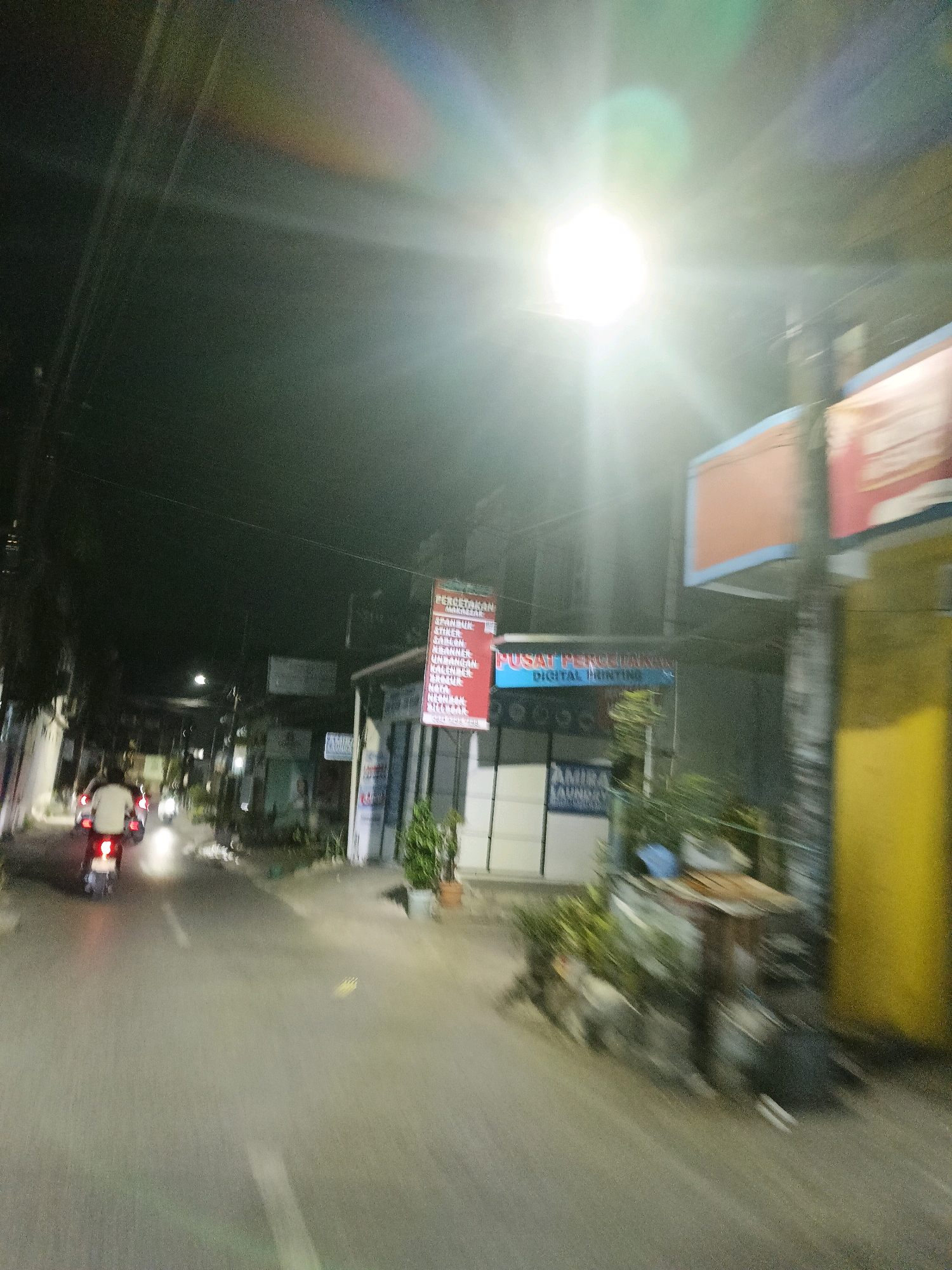 A night-time, motion-blurred, street-level photograph taken from the perspective of someone riding a motorcycle in Makassar, Indonesia. The image captures a residential or commercial street with moderate lighting from a bright overhead streetlamp causing lens flare. On the right side of the road, several signs are visible. One red sign reads "PERCETAKAN" followed by a list of printing services including "SPANDUK", "STIKER", "CARLON", "KANTO", "UNDANGAN", "KOLER", "KALENDER", "NOTA", "KWITANSI", and "POSTER". Below this, a blue sign states "PUSAT PERCETAKAN DIGITAL PRINTING". Further down, a sign for "AMIRA LAUNDRY" is visible. A bright yellow column is also present on the right. On the left side of the road, a few buildings and trees are partially visible. Two motorcycles are traveling away from the camera, with the rider of the leading motorcycle wearing a white shirt. The road surface is asphalt with a white line marking the center. The overall impression is that of a busy, commercially active street in Makassar at night.