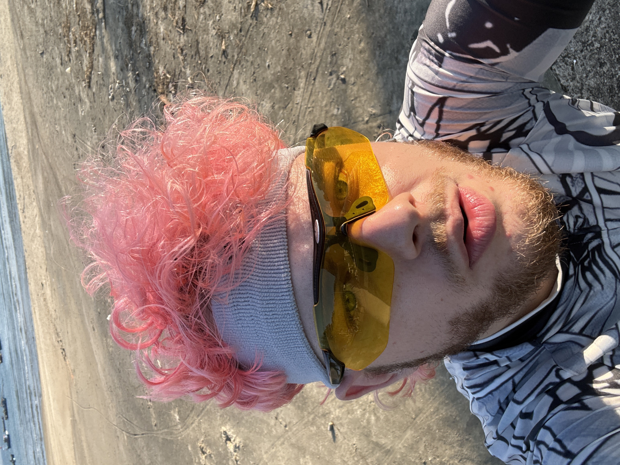 A person is lying on their back on a sandy beach. The person has pink curly hair, a gray headband, and yellow sunglasses. They are wearing a black and white patterned garment. The beach is sandy and the sea can be seen in the background. The location is likely a beach in Đà Nẵng, Vietnam, given the setting. The subject is posing for a selfie.