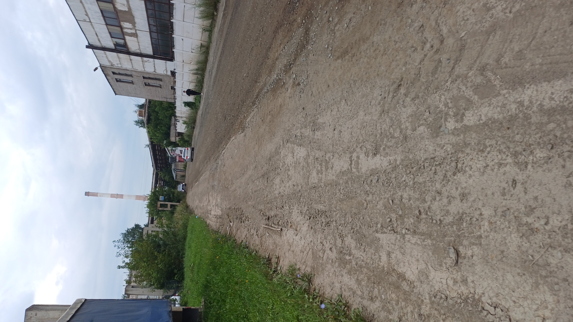 The image shows a dirt road in an industrial area. A tall, striped smokestack is visible in the background, along with a long, light-colored building. There are a few cars on the road, and one person walking away from the camera. The area is overgrown with grass and trees. The scene is likely located in Izhevsk, Russia, given the presence of industrial infrastructure and a smokestack typical of such areas.