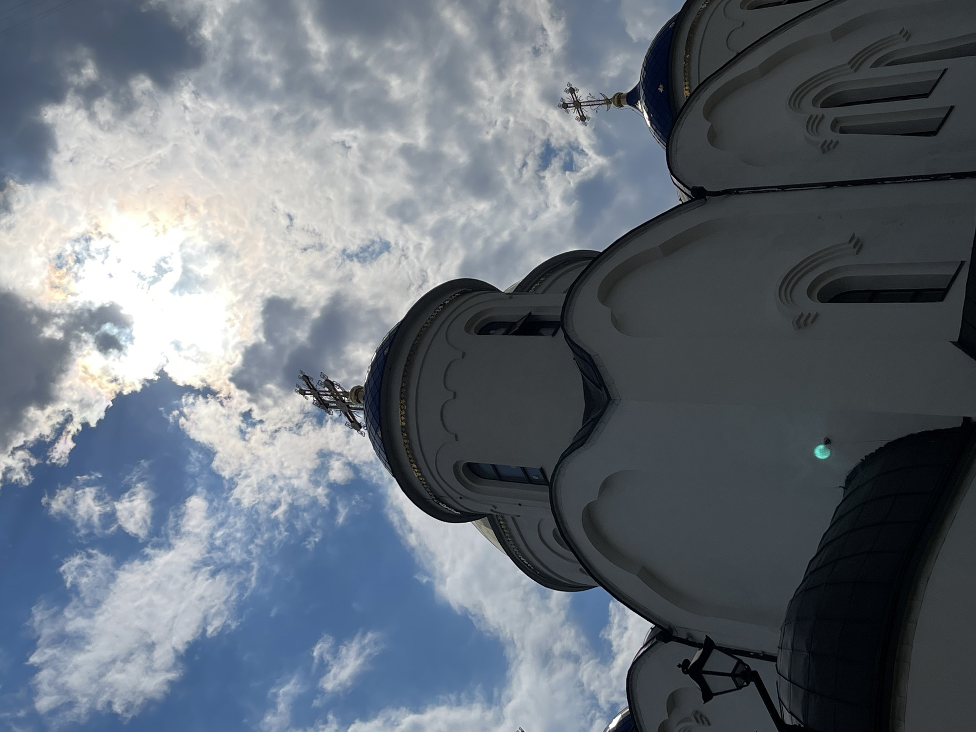 The image shows a low-angle, wide shot of a white church with blue domes against a cloudy sky. The sun is visible through the clouds, creating a bright halo effect. The church has multiple domes, each topped with a cross. The architecture appears to be Orthodox Christian. There is a streetlamp attached to the side of the building. The church's location in Moscow, Russia is not specified in the image.