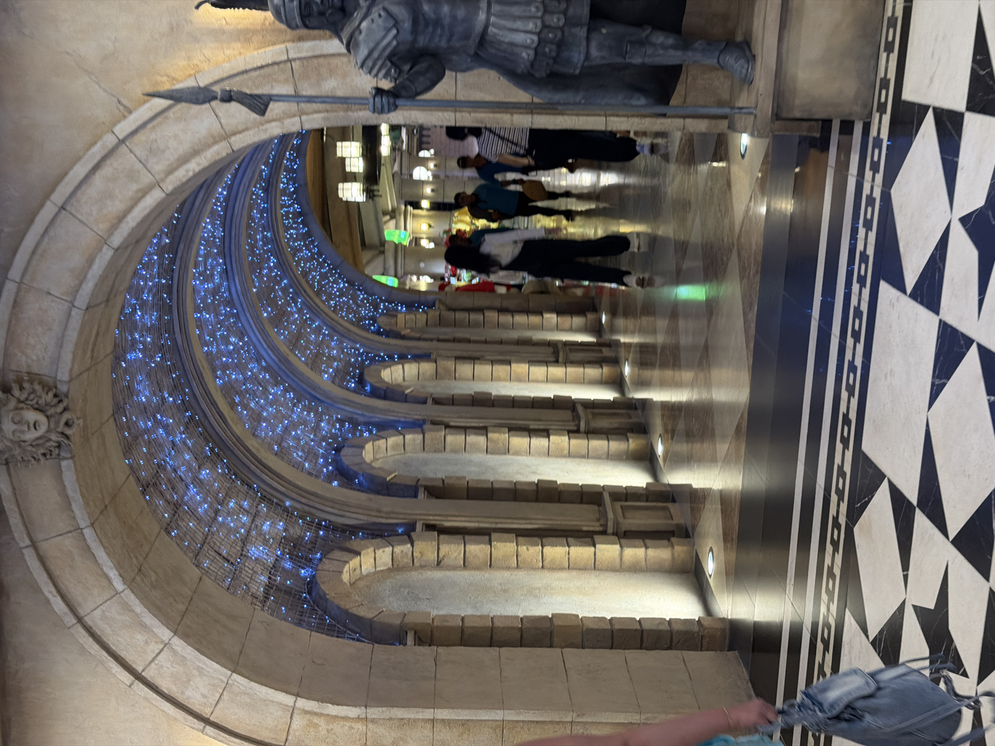 The image depicts an interior view of a shopping mall or entertainment venue, likely located in Đà Nẵng, Vietnam. The scene features a grand archway leading into a long corridor illuminated by blue fairy lights overhead. The architecture is reminiscent of Roman or classical styles, with stone arches and decorative elements. To the right, a large, dark statue of a warrior in armor stands guard, holding a spear.
Several individuals are visible walking through the corridor. In the foreground, a person is walking away from the camera, carrying a light blue backpack. Further down the corridor, a group of people are walking towards the viewer. One person in the middle ground is wearing black overalls and a white shirt, another is wearing a blue shirt, and others are less distinctly visible.
The floor is decorated with a geometric pattern of black and white tiles, with a black border featuring a repeating square design. The lighting within the corridor is a mix of ambient light from the overhead decorations, spotlights on the pillars, and what appears to be ceiling lights further down the hallway, casting reflections on the polished floor. The overall impression is one of an indoor recreational space with a theatrical or themed design.