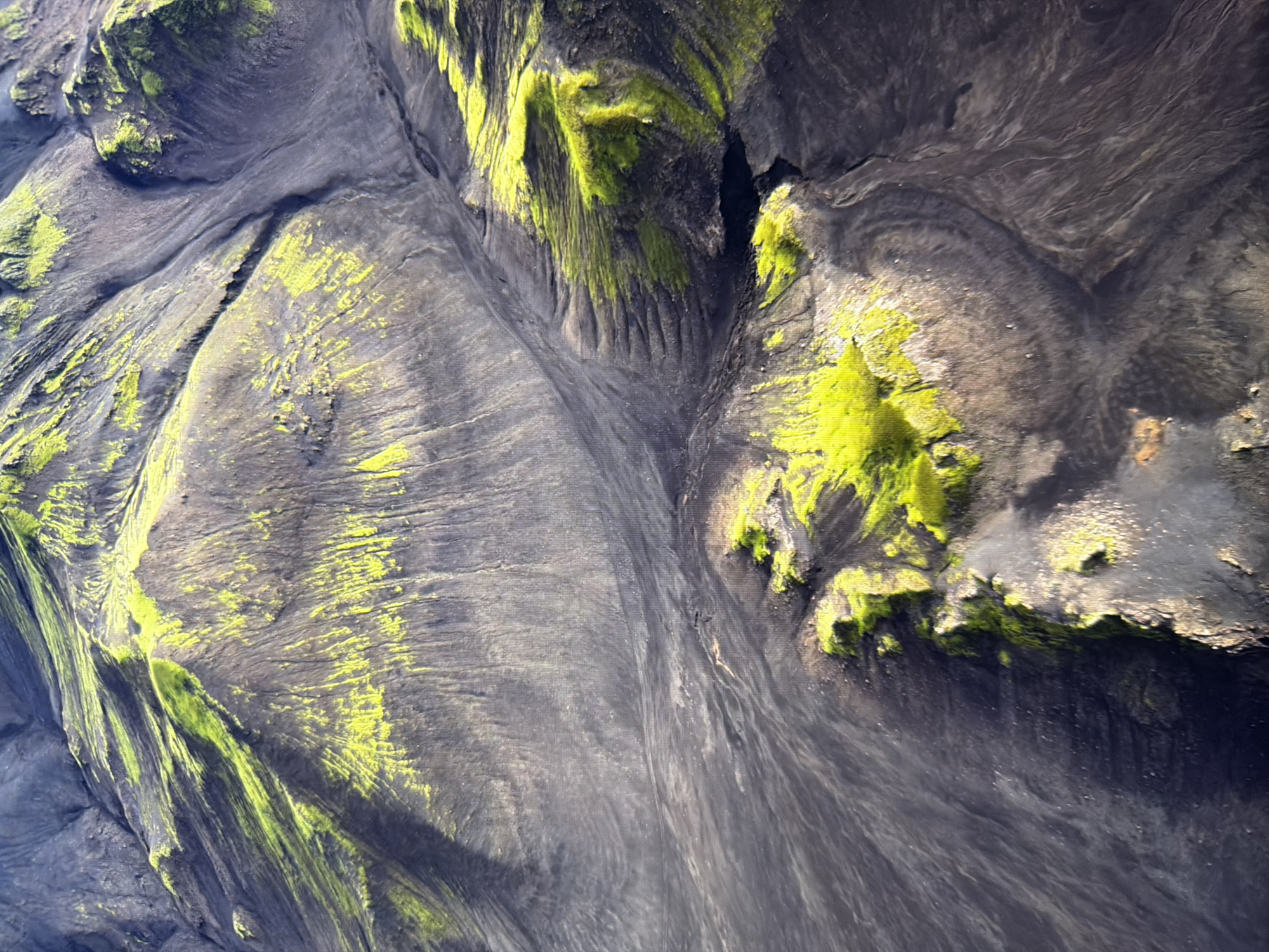 This is an aerial photograph of a barren, mountainous landscape. The terrain is predominantly dark gray and appears to be composed of volcanic ash or rock. Patches of vibrant green moss or vegetation are scattered across the slopes, creating a stark contrast with the dark ground. The landscape is characterized by erosion patterns, with visible gullies and ridges formed by water or wind. The lighting suggests it is daytime, with sunlight illuminating some areas and casting shadows on others, highlighting the topographical features. There are no visible signs of human presence, buildings, or city structures. Therefore, this image is not of Kyiv, Ukraine.
