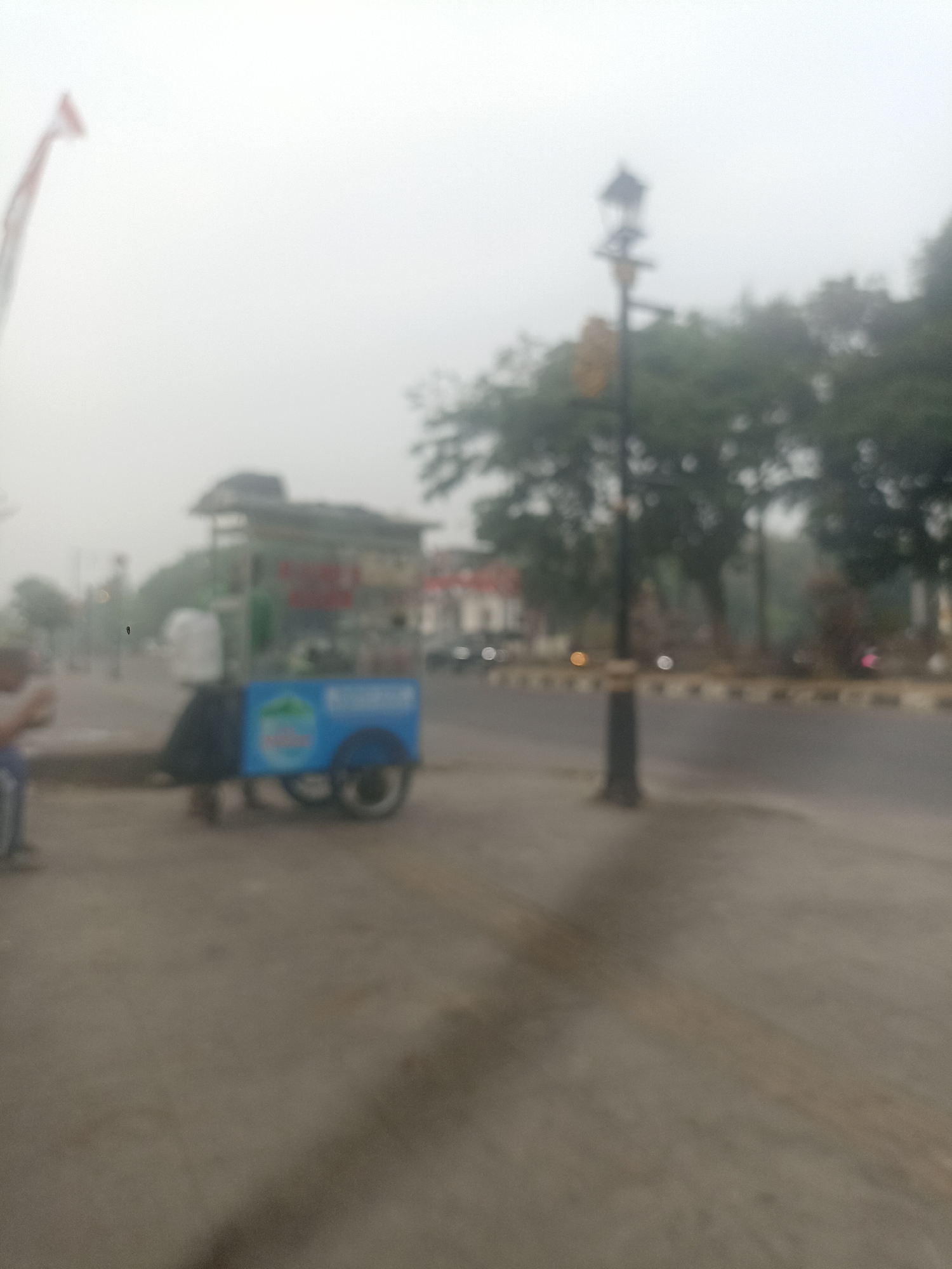 The image, captured in the city of Pamekaran, Indonesia, depicts a slightly out-of-focus street scene on a hazy day. In the foreground, a two-wheeled cart with a blue body and a white container on top is parked. The cart has some signage on its side, but it is illegible due to the blurriness. To the left of the cart, a person is partially visible, seated and holding something, possibly a drink. Further into the background, a street lamp with a distinctive square-shaped top stands near a road. Trees line the opposite side of the road, and a building with a red and white facade is visible in the distance. An Indonesian flag is also faintly visible on a pole to the far left of the image. The overall atmosphere suggests a quiet, possibly early morning or late afternoon setting, with subdued lighting and limited visibility due to fog or haze.