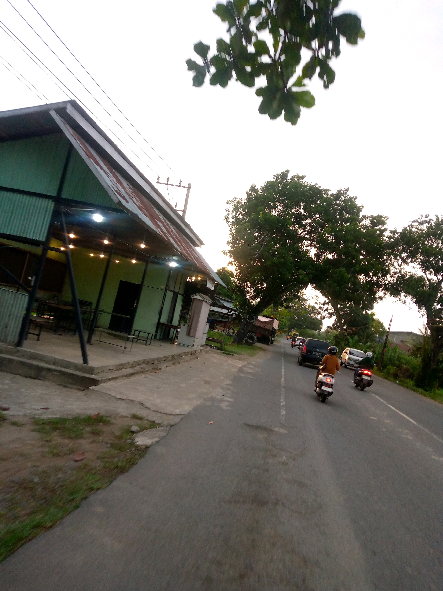 The image captures a street scene in Keudeteupinpunti, Indonesia, with a lean-to-style building on the left and a road with traffic on the right. The building, painted light green with a corrugated metal roof, appears to be an open-air establishment with tables and chairs set up on a concrete platform. String lights are visible illuminating the interior. A few utility poles and power lines run overhead.
The road is paved and shows signs of wear. Traffic includes several motorbikes and a black SUV. Two individuals are riding motorbikes in the foreground, one with a person wearing a light brown jacket. On the right side of the road, a large tree with dense foliage arches over the road, partially obscuring the sky. Another truck with a red trailer is parked on the left side of the road further down. The overall lighting suggests it is late afternoon or early evening.