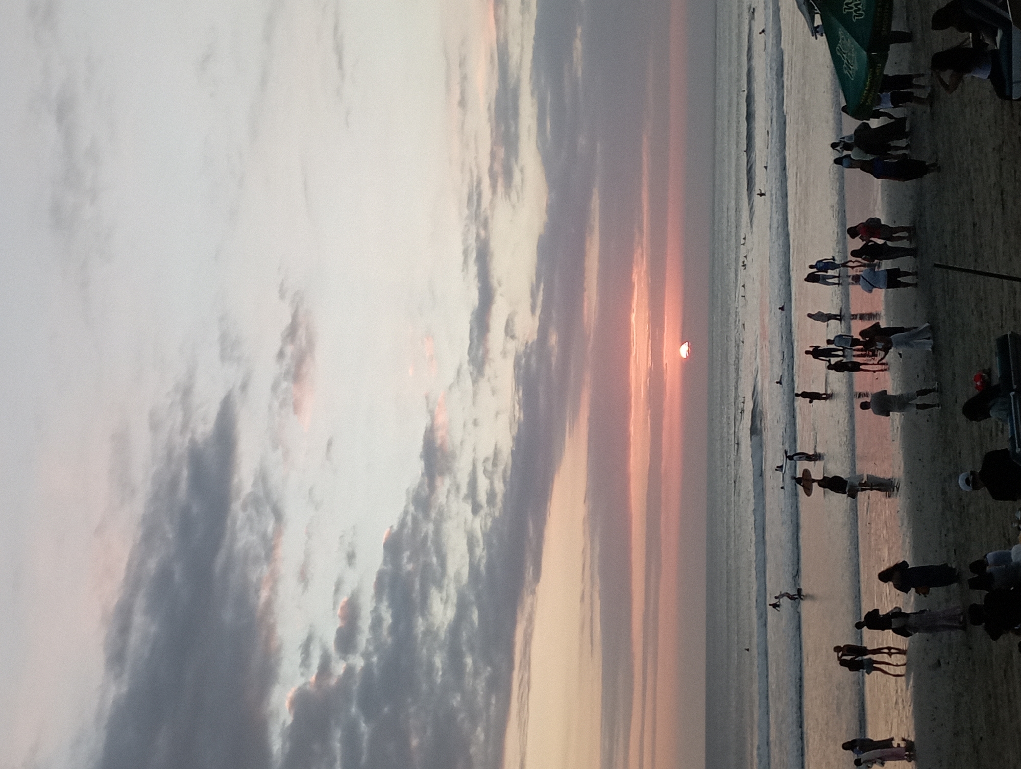 At sunset on Kuta Beach, Indonesia, a crowd gathers as the sun, a vibrant orange orb, dips below the horizon. The sky is painted with hues of pink, orange, and gray, with clouds streaking across the sky. The ocean is calm, with gentle waves lapping at the shore. People are scattered across the beach, some wading in the water, others standing on the sand, and some with surfboards. In the foreground, two green umbrellas provide shade for beachgoers. The overall atmosphere is one of relaxation and enjoyment.