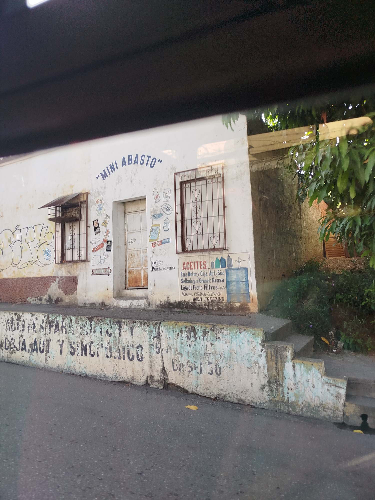The image shows a small building with the words "MINI ABASTO" painted on its facade. The building appears to be a shop or convenience store, as indicated by the word "ABAsto," which means grocery store in Spanish. Various items are displayed on the wall, including what appear to be labels for lubricants and automotive fluids. There are also some decorative elements and graffiti on the building. The building is situated on a street, with a concrete barrier in the foreground. The surrounding environment suggests an urban or semi-urban setting, with trees and vegetation visible in the background. The overall impression is of a modest commercial establishment in a local neighborhood.
