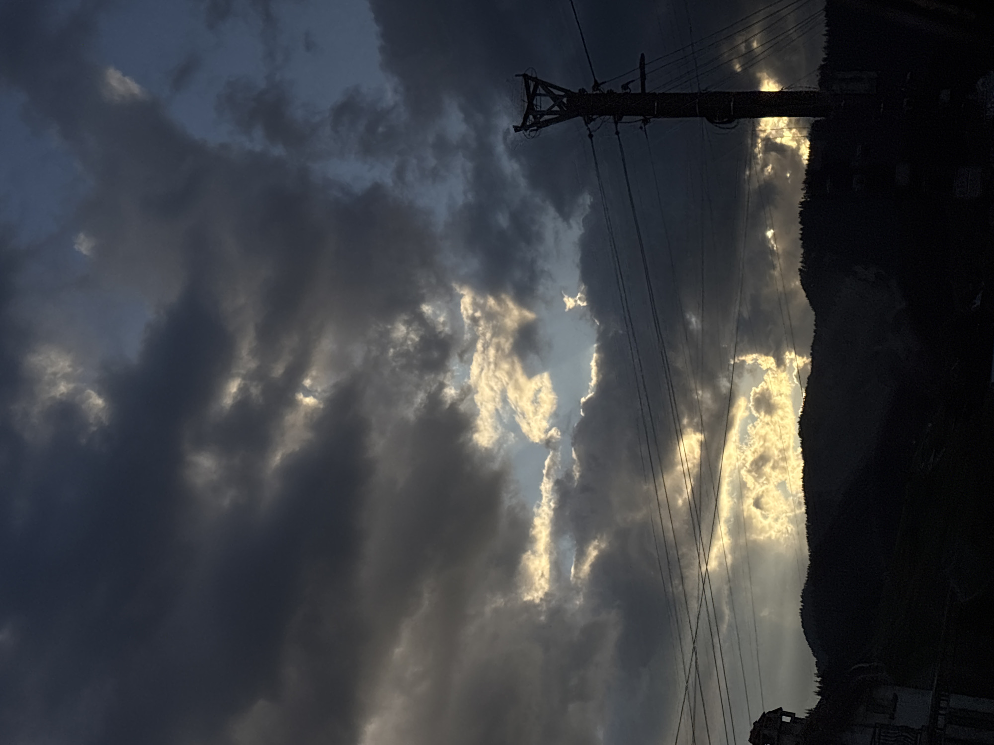 The image shows a view of the sky over Borino, Bulgaria. The sky is filled with dark, ominous clouds, but there are a few breaks in the clouds that allow sunlight to shine through. In the foreground, there is a telephone pole. To the right, there is a dark, tree-covered mountain. At the bottom right corner, there are a few buildings.