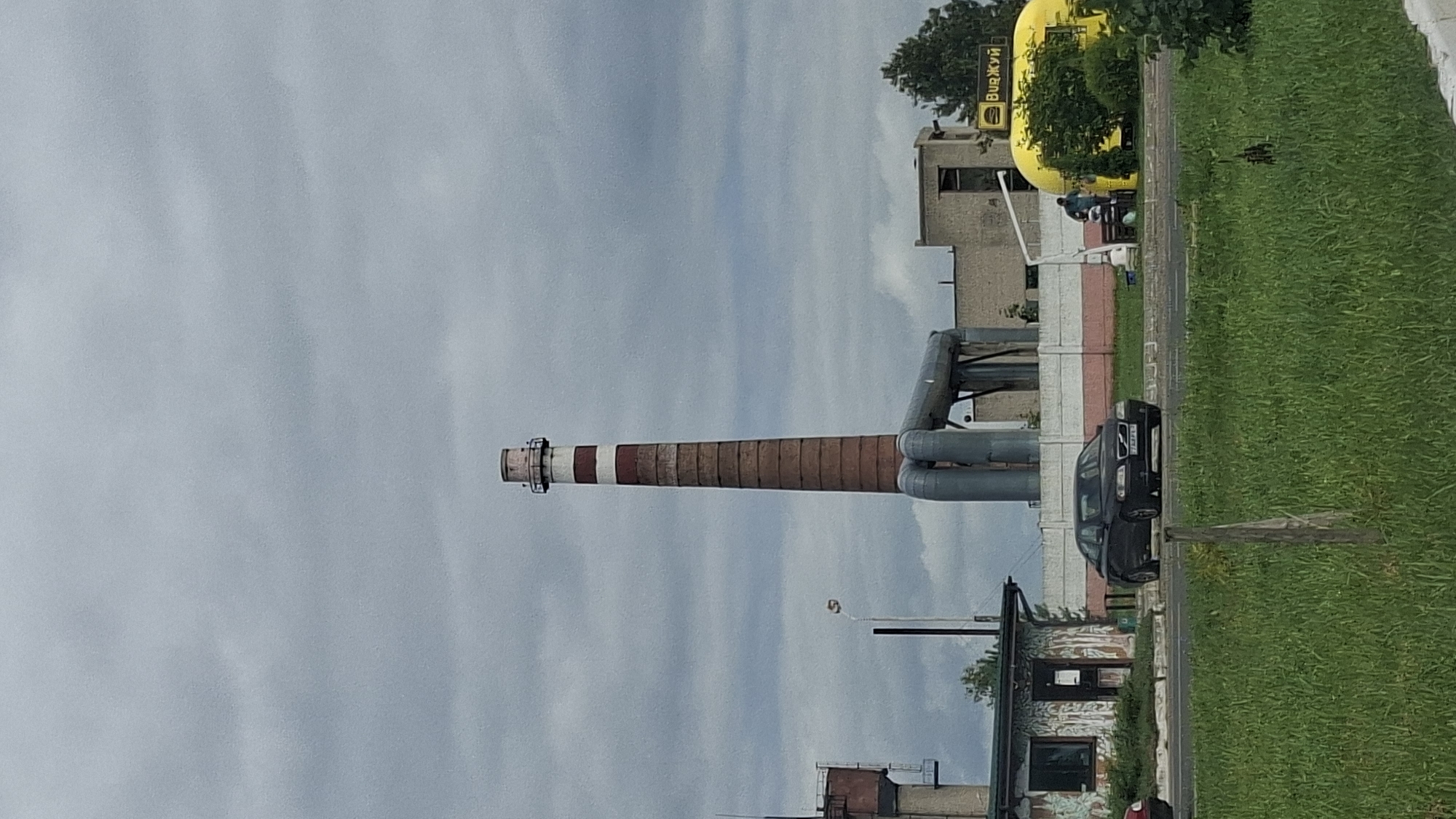 The image depicts an industrial area on an overcast day. In the center of the frame stands a tall, brick smokestack with red and white bands, supported by large, grey pipes. To the left of the smokestack, a water tower is visible. In the foreground, a dark sedan is parked on a paved surface. Adjacent to the sedan is a small, weathered building with a green roof and graffiti on its walls. To the right of the sedan, a bright yellow, possibly food-related structure is present, with a sign above it that reads "ВидЖУЙ" in Cyrillic script. Two individuals are visible near the yellow structure, one standing and another sitting. The ground is covered in lush green grass. The location is identified as Myadzyel, Belarus.