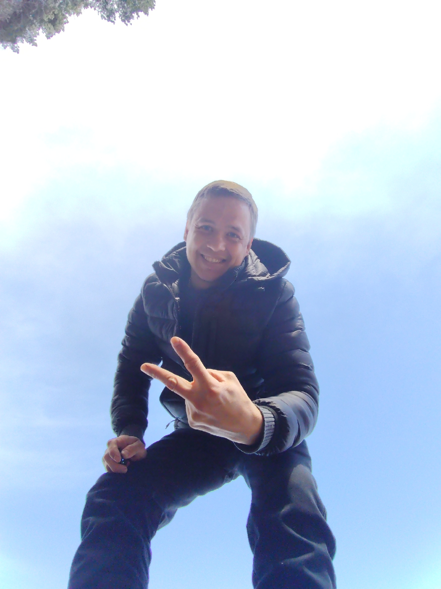 An adult man is captured in a low-angle shot, smiling broadly and looking directly at the camera. He is making a peace sign (V-sign) with his right hand, held at chest level. The man is wearing a dark, possibly black, puffer jacket and dark pants. His expression conveys happiness and friendliness.
The setting is outdoors, with the man silhouetted against a bright, expansive sky. The sky is predominantly clear blue towards the bottom, transitioning to a lighter, almost white tone towards the top, with hints of faint cloud cover. In the upper left corner, a small section of dark green foliage, likely from tree branches, is visible, suggesting an environment with natural elements. The bright light indicates it is daytime with good weather. There is no visible text or specific architectural details to confirm the location as Chaikovsky, Russia, but the scene is consistent with an outdoor, possibly park-like, environment. The unusual low camera angle emphasizes the man's presence against the sky.