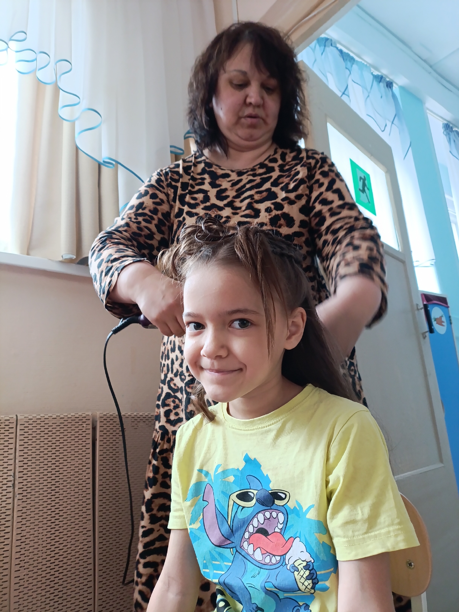 In Chaikovsky, Russia, an interior scene features a woman with dark brown hair styled in loose waves, wearing a brown leopard print dress, standing behind a seated young girl. The girl has light brown hair, partially styled and held back, a smiling face, and is wearing a yellow T-shirt with a cartoon character printed on it. The woman is styling the girl's hair with a heated styling tool. A radiator is visible to the left, and in the background, there are windows covered with light curtains, a door with a green exit sign, and a blue decorative element.