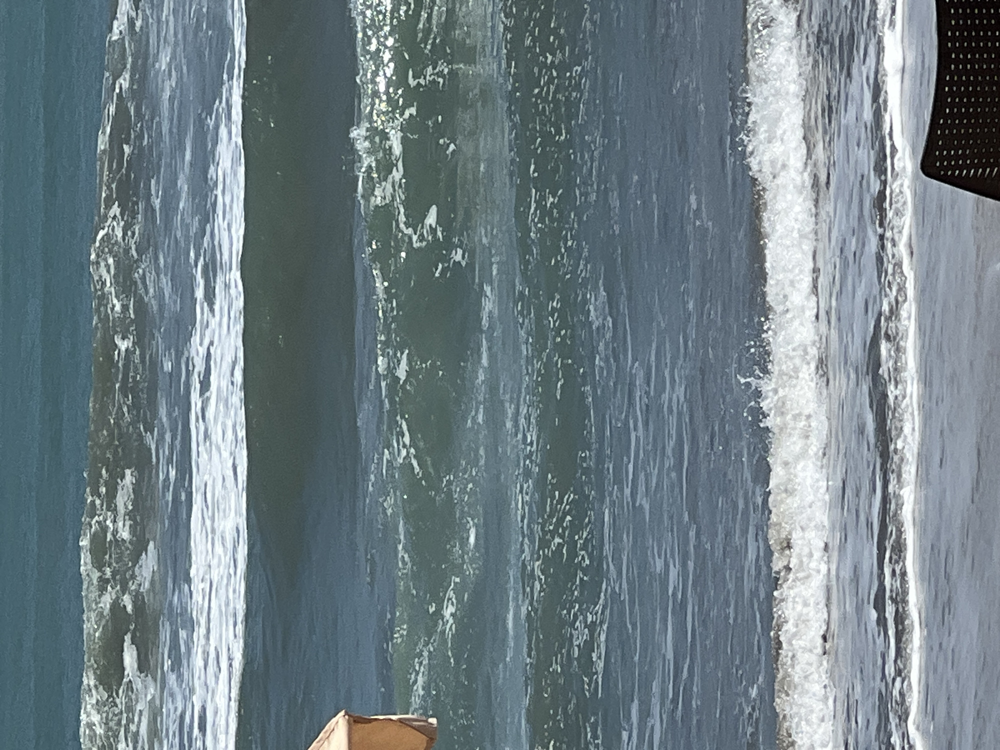 This is a medium-shot, eye-level photograph of ocean waves near the shore. The ocean is a deep blue-green, with multiple waves rolling in. The closest wave to the viewer is breaking, creating white foam and spray. In the background, there are several other waves, some with whitecaps. A small portion of an orange umbrella is visible on the left side of the frame, and a dark-colored chair is visible on the bottom right. The overall impression is one of a calm, sunny day at the beach. The location is the coast of Kuta, Indonesia, a popular tourist destination known for its beaches and surf.