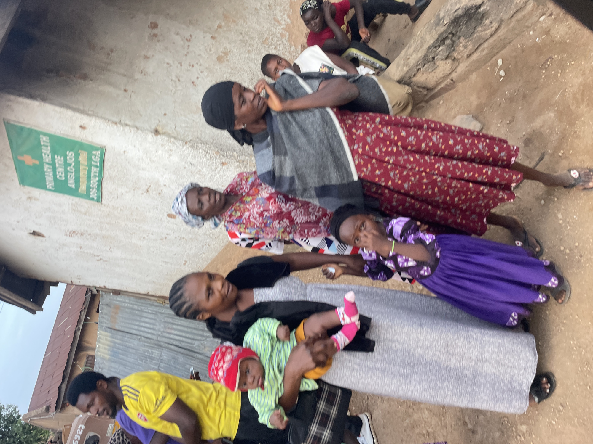 The image shows a group of people standing and sitting outside a building in Jos, Nigeria. A sign reading "PRIMARY HEALTH CENTRE ANGLO-JOS JOS-SOUTH LGA" is visible on the building. Several adults and children are present. One woman is holding a baby, while another child is standing next to her. Some people are sitting on a concrete structure. The environment appears to be a dusty outdoor area.