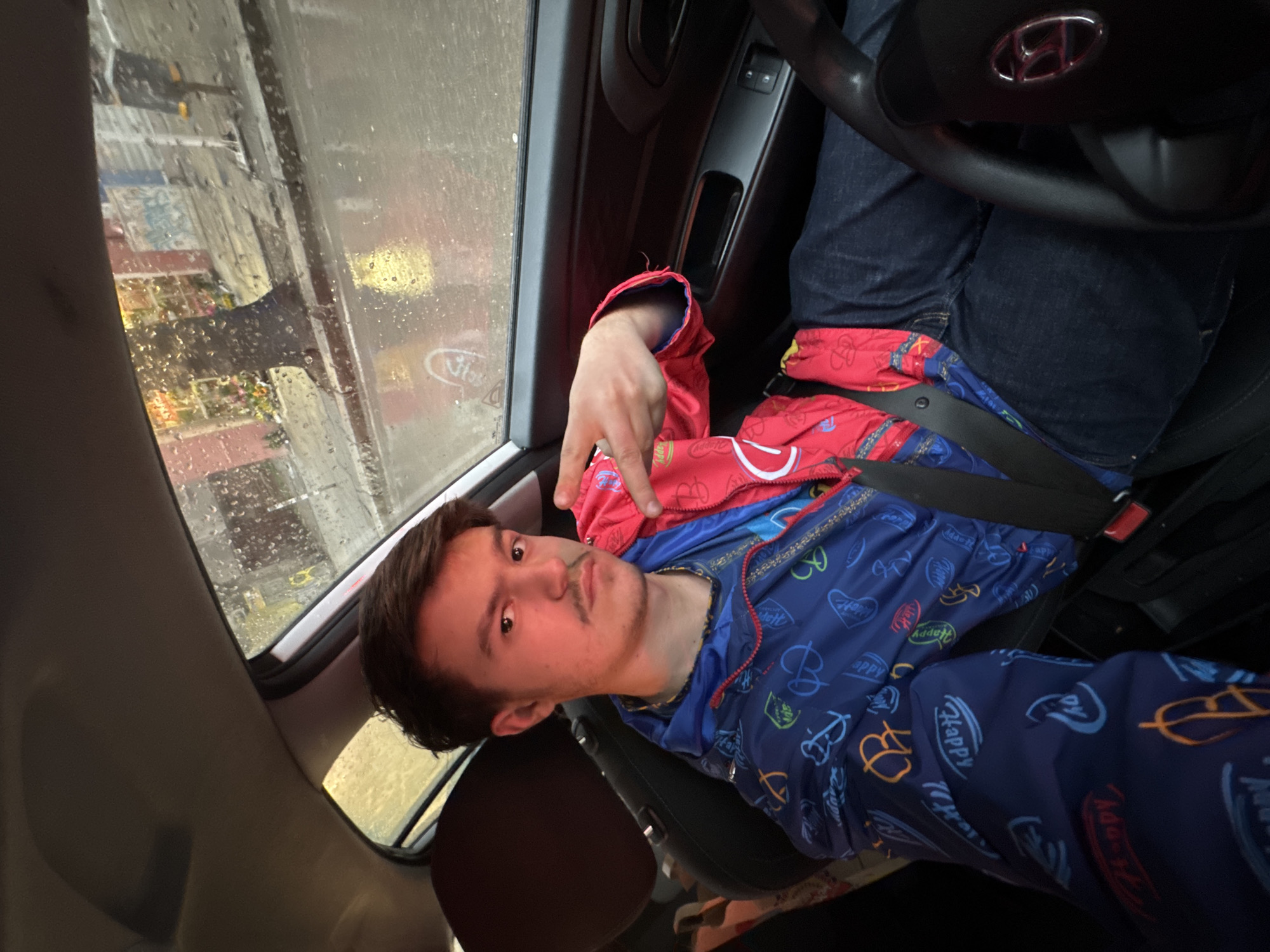The image shows a young man with dark hair and a mustache sitting in the driver's seat of a car. He is wearing a blue jacket with cartoon-like designs and a red lining, and a seatbelt is fastened across his body. His left hand is raised near his face, making a gesture. The interior of the car is visible, including the steering wheel with a brand emblem and the dashboard. Through the car window, a blurred cityscape is visible, with rain droplets on the glass. The location is Sofia, Bulgaria, as evidenced by the Cyrillic script and architectural style in the city seen through the car window.