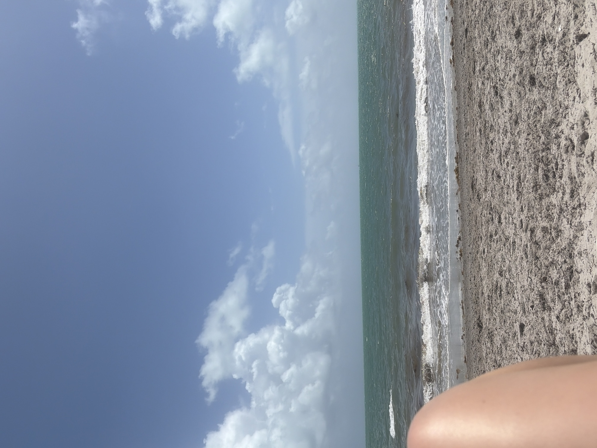 The image shows a beach scene, likely on Key Biscayne in the United States. The sky is mostly blue with scattered white clouds. The ocean is turquoise-green with small white waves breaking onto the shore. The beach consists of sand that is a light gray color. In the lower right corner, a portion of a human leg is visible.