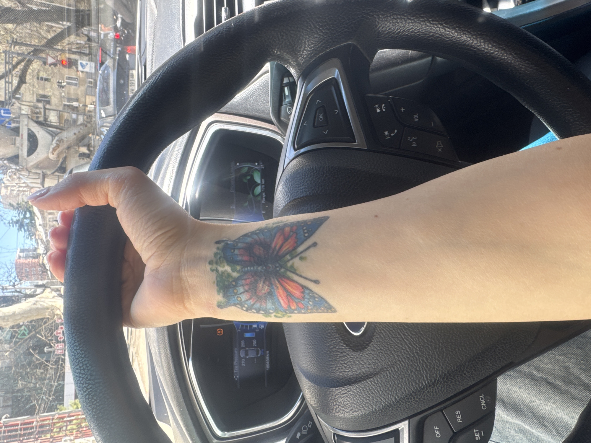 The image shows the interior of a car, specifically focusing on the driver's side. A person's left hand and forearm are visible, gripping the steering wheel. The arm has a butterfly tattoo on the wrist. The car's dashboard and instrument panel are partially visible, showing readings like tire pressure and remaining fuel range. Through the car's front window, a cityscape is seen, recognizable as Tbilisi, Georgia, due to the architectural style and street signs. Traffic and buildings are visible in the background.