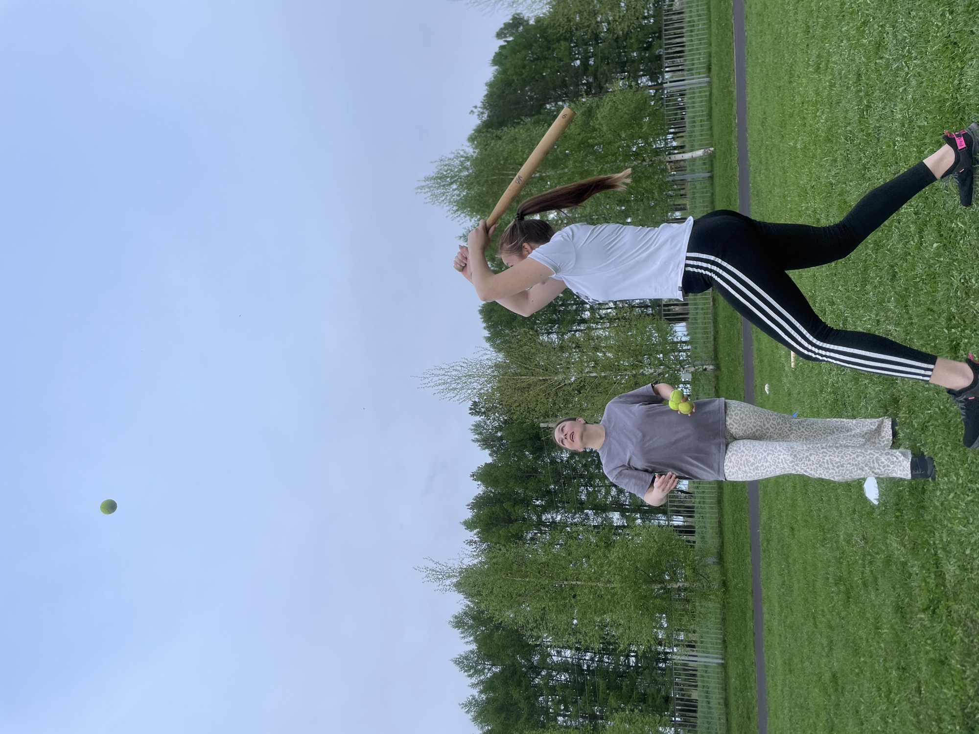 Two females are in a field in Подшивалово, Russia. One is holding a baseball bat, swinging it. The other is holding two tennis balls. The field is grassy with trees and a fence in the background. The sky is overcast.