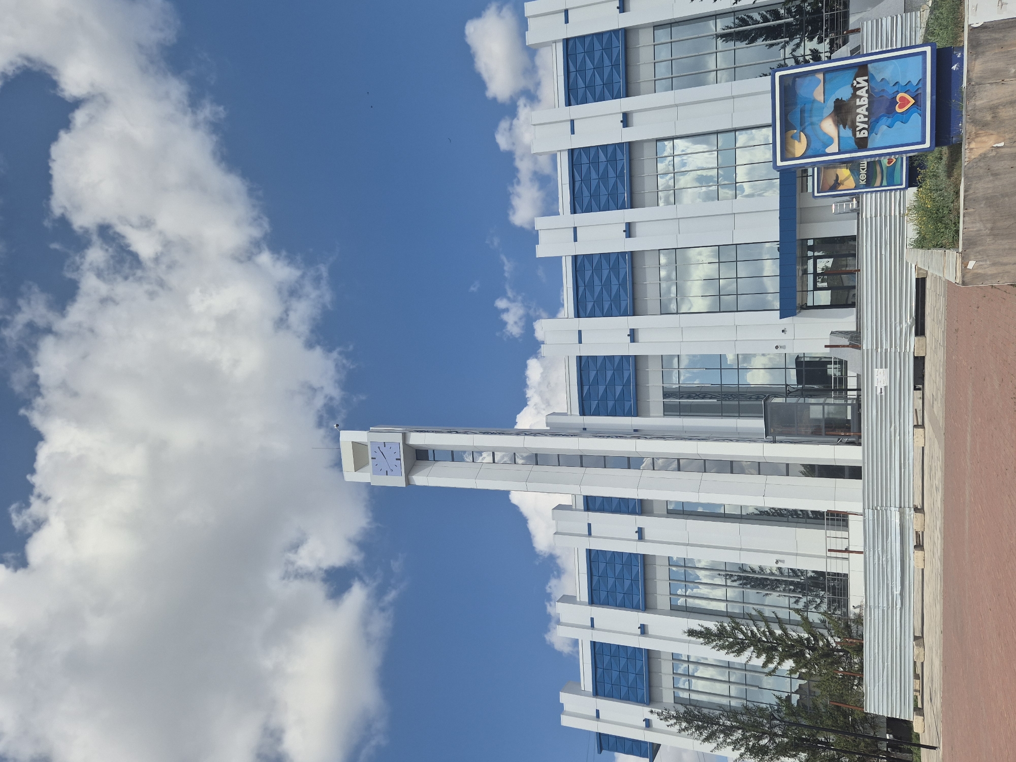 The image captures the modern architecture of the Kokshetau train station in Kokshetau, Kazakhstan, under a bright blue sky with scattered white clouds. The station features a tall, white clock tower with a square clock face displaying Roman numerals, centrally positioned above a broad, white facade accented with blue decorative panels. Large windows reflect the sky and surrounding trees, giving the building a clean and contemporary appearance. In the foreground, a red brick sidewalk leads to the station entrance, with a temporary corrugated metal barrier and some greenery visible. Two colorful signs are placed near the entrance, one prominently displaying the word "БУРАБАЙ" (Burabay), a popular tourist destination near Kokshetau. The scene is devoid of people, suggesting a quiet moment at the station. The overall impression is one of a well-maintained and functional transportation hub in a visually appealing urban setting.