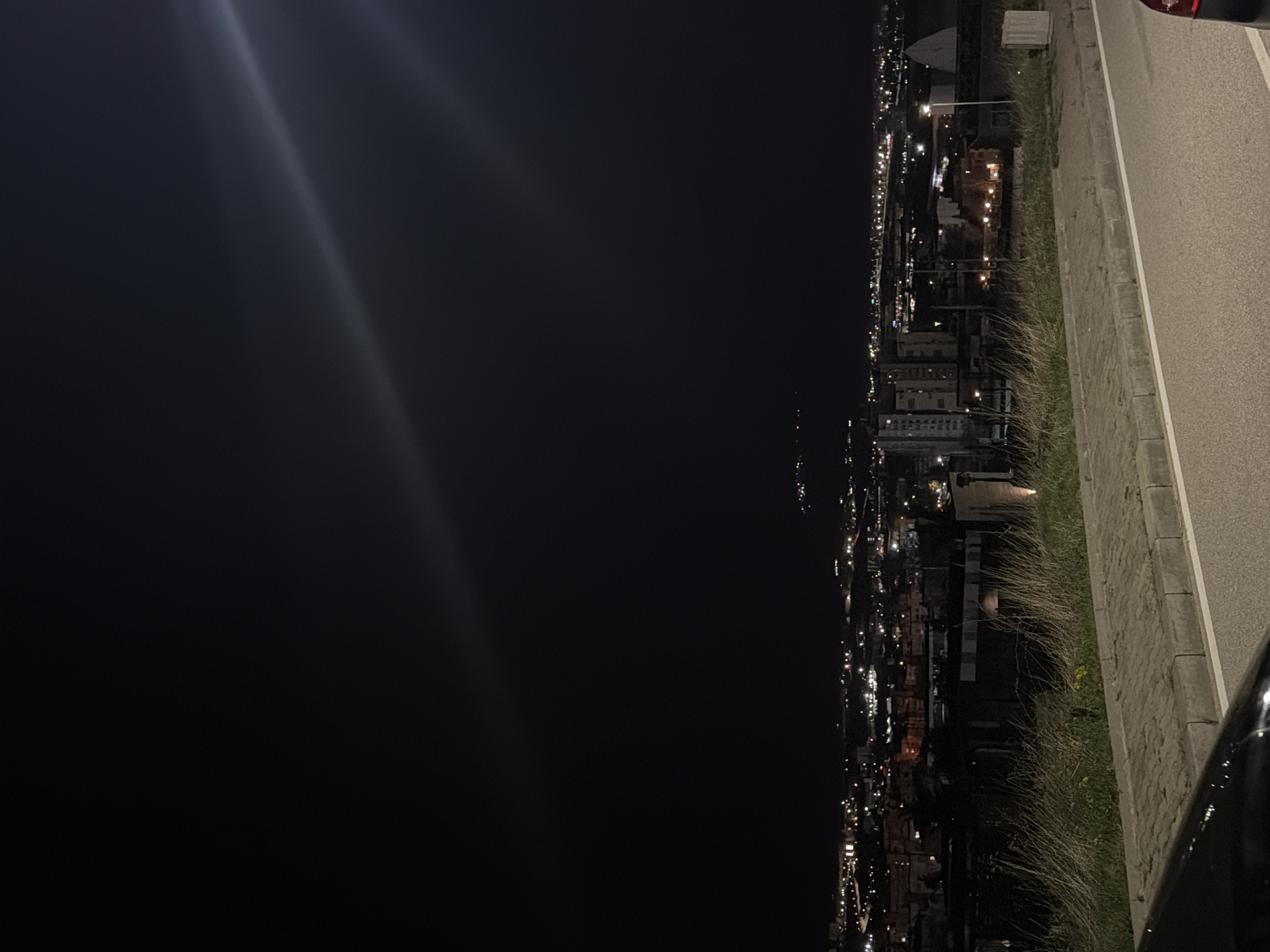 Captured at night, the image presents a panorama of Blagoevgrad, Bulgaria, viewed from an elevated roadside perspective. The left side of the composition is dominated by the night sky, while the cityscape, illuminated by various lights, stretches along the right. A portion of a roadway, bordered by a grassy verge and a pavement, is visible, suggesting the viewpoint is from a parked vehicle, with parts of a car's body visible. The city below shows a dense arrangement of buildings and streetlights, creating a grid-like pattern of light against the dark background. No people are visible in the frame.