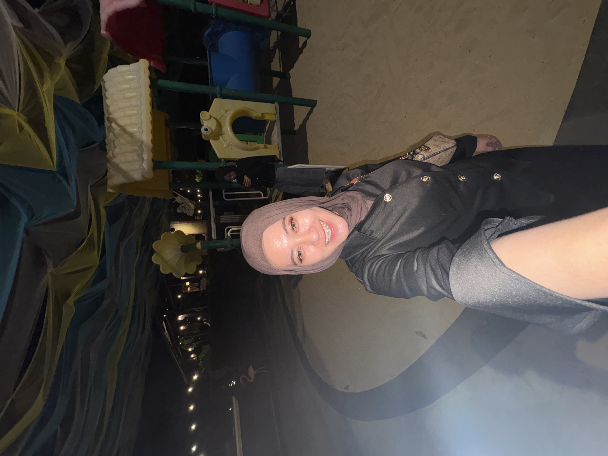 A selfie is taken by a woman in a brown hijab and a black coat. She is smiling and has braces. Behind her is a playground with colorful slides and structures. The playground is covered with a canopy of blue and yellow fabric. The scene appears to be in a public area, possibly a park, at night. The location is identified as Bulusan, Indonesia.