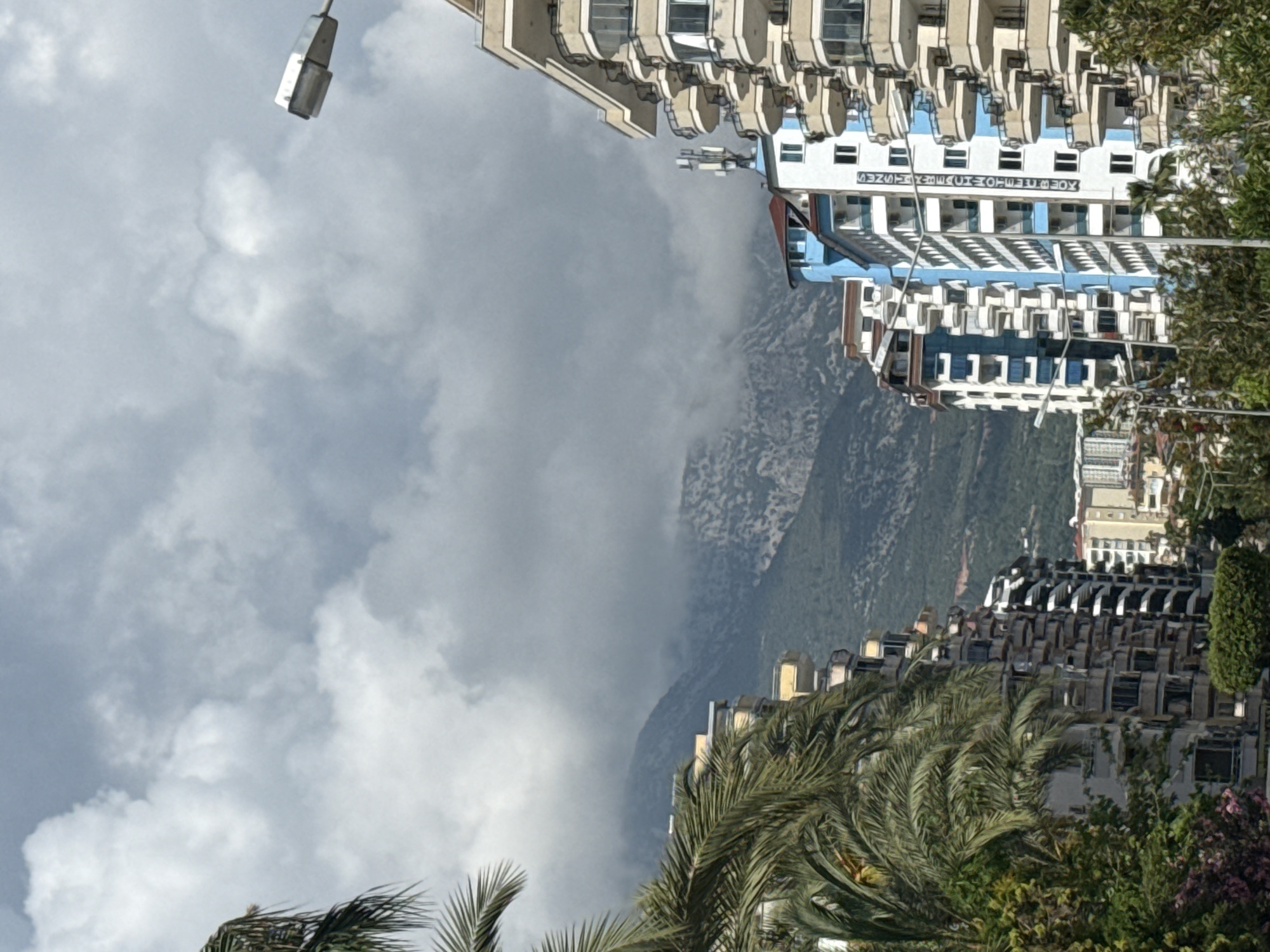 The image depicts a scene in an unknown city, featuring a view of the sky with varying cloud cover. Two buildings are visible on the left and right sides of the image. On the left side, a taller building with white balconies, and on the right side, a building with black balconies. In the middle ground, a mountain is partially obscured by clouds. Palm trees and other greenery are visible near the bottom of the image, suggesting a tropical or subtropical environment. A street light is on the upper left side of the image.