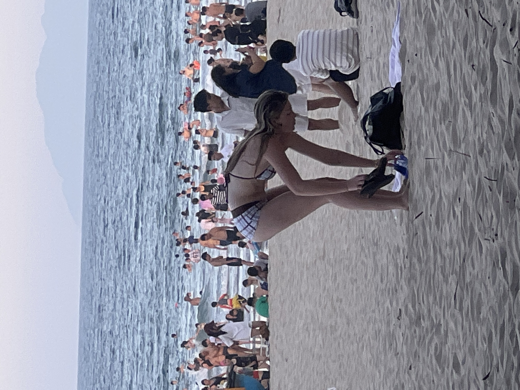 The image depicts a crowded beach scene on a cloudy day in Da Nang, Vietnam. The ocean is calm with gentle waves and many people are swimming and wading in the water. On the sandy beach, a young woman in a checkered bikini is bending down, holding a pair of black flip-flops. Beside her is a black beach bag and a white towel. In the background, a diverse group of individuals are visible, some walking on the shore, others interacting or relaxing. The horizon is marked by hazy mountains. The overall atmosphere suggests a popular tourist destination during a day with overcast skies.