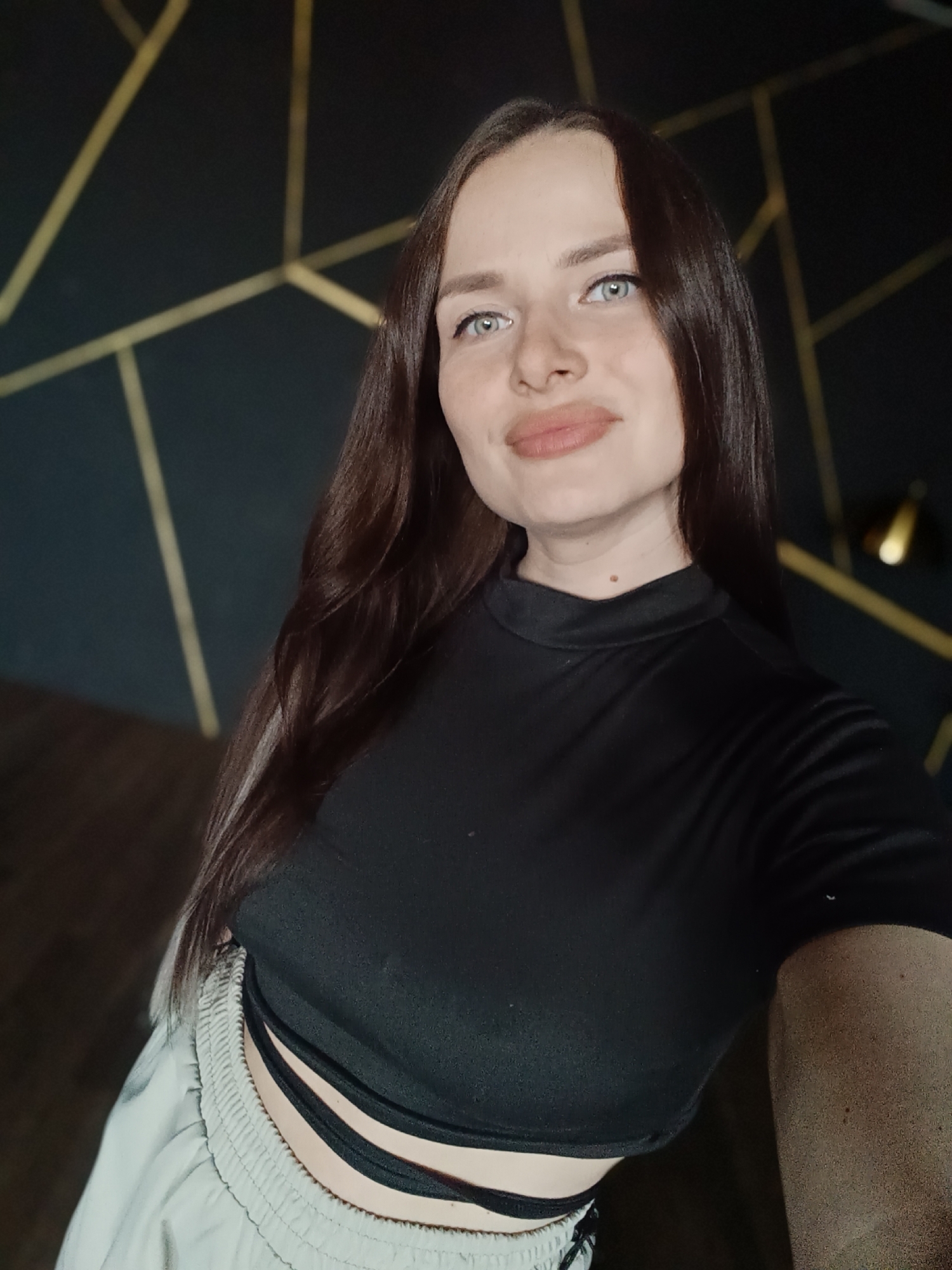 A medium shot depicts a fair-skinned young woman with long, straight, dark brown hair. She faces forward, glancing slightly to her right. Her eyes are light blue. She has full lips painted with a matte, nude color. She is wearing a short-sleeved black mock turtleneck shirt with an exposed midriff. Her bottoms appear to be light-colored, possibly gray, sweatpants. The background is a dark wall with a gold geometric pattern of intersecting lines. To the right, a gold-colored object, possibly a lamp, is partially visible. The location is Unknown, Unknown.
