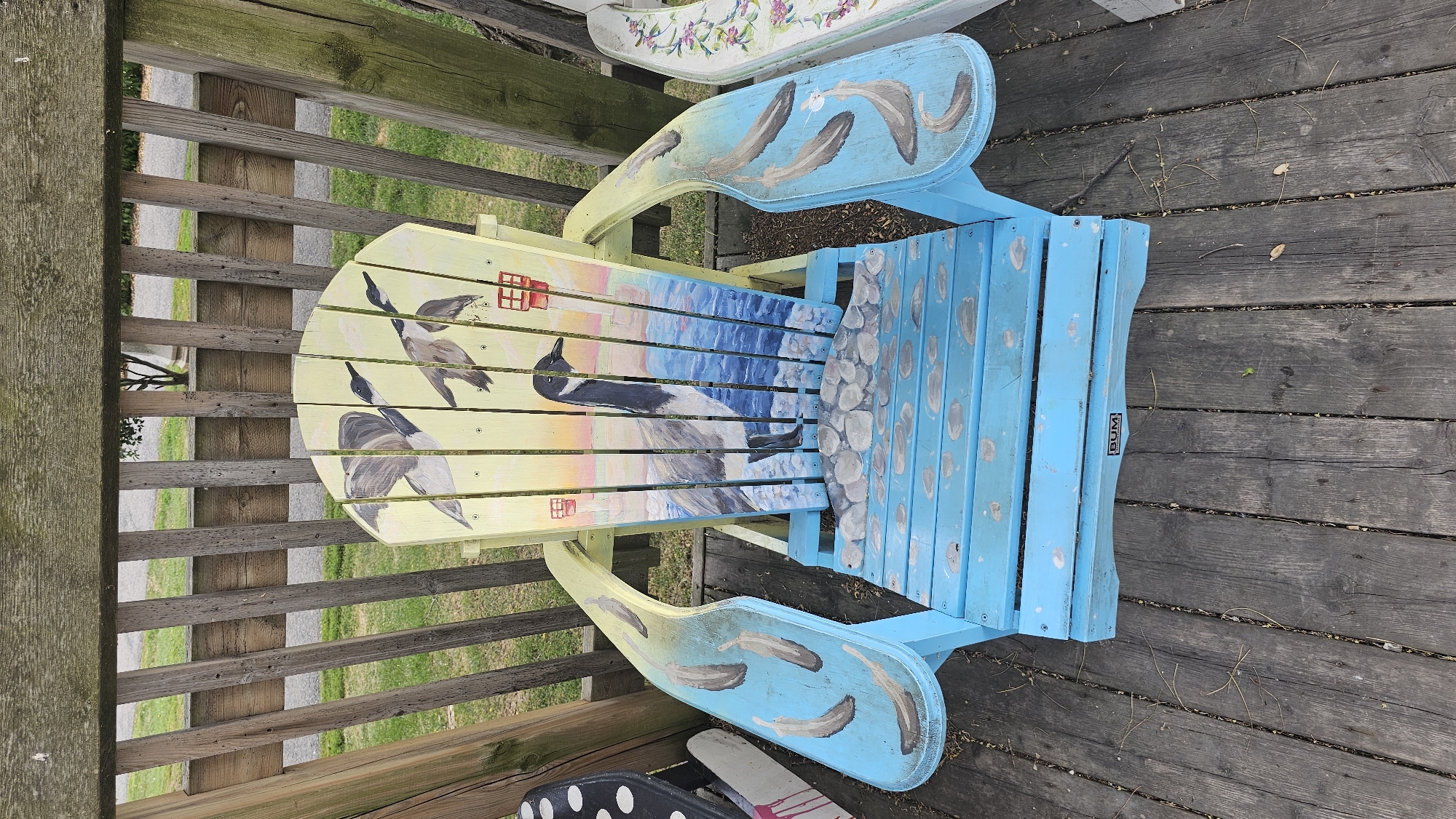 An overhead, rotated view shows a hand-painted Adirondack chair on a weathered wooden deck. The chair is adorned with a vibrant mural depicting a coastal scene. The backrest and seat are painted in a gradient of yellow and blue, transitioning to represent a sunset over the water. Two Canadian geese are painted in flight on the yellow portion of the backrest, with a small red house also visible. The seat features a depiction of water and rocks. The armrests are painted blue with abstract feather-like designs. The wooden deck beneath the chair is composed of dark, aged planks. The background reveals a wooden fence and green grass, suggesting an outdoor setting. A small black and white label with the word "BUM" is visible on the side of the chair. Another hand-painted chair with a floral design is partially visible behind it. The lighting suggests it is daytime.