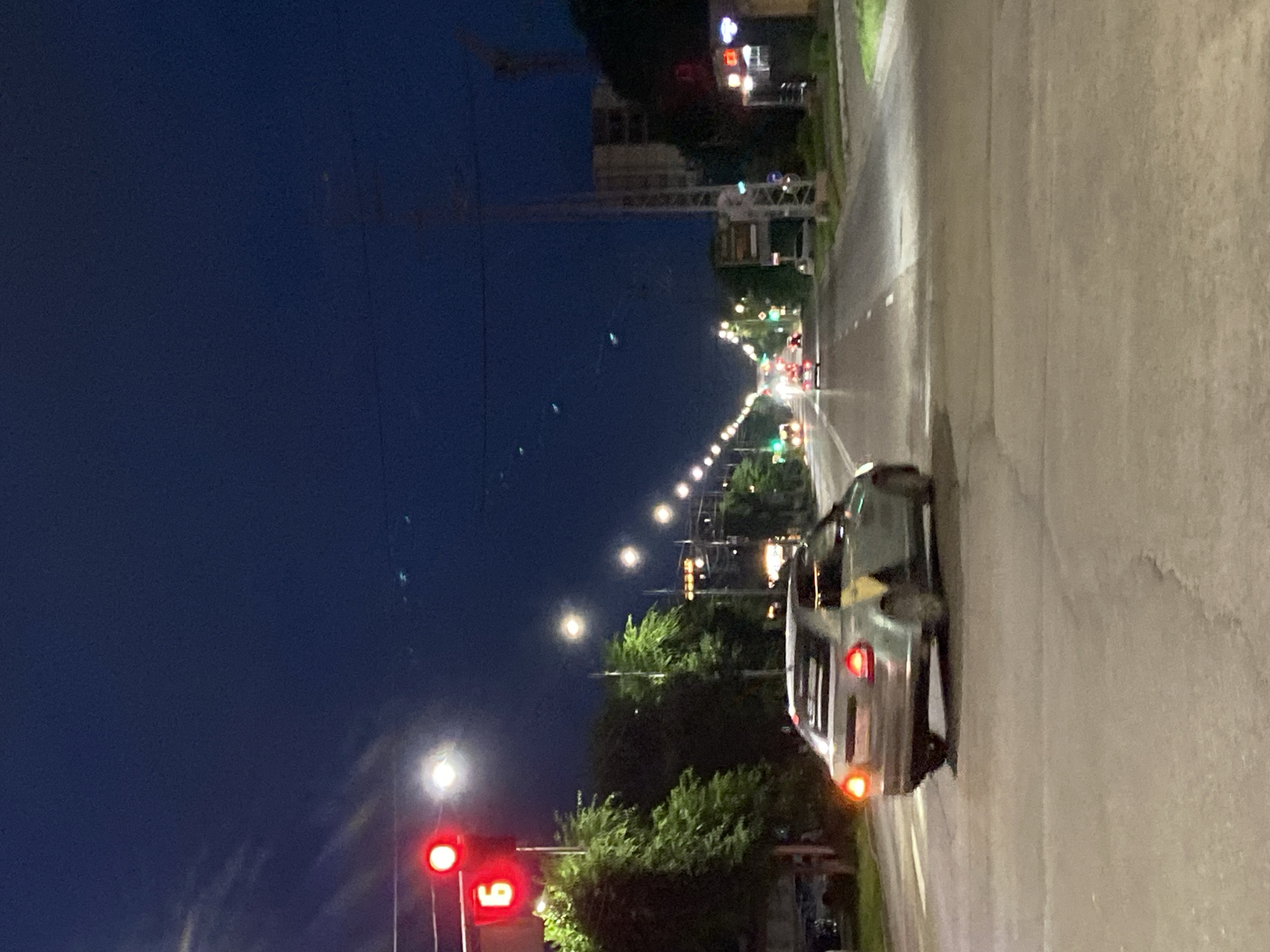 The image depicts a street scene at dusk in Blagoveshchensk, Russia. A silver sedan is captured mid-turn on the right side of the road. The car's brake lights are illuminated, indicating it is decelerating or stopped. In the background, the street is lined with trees and streetlights, with other vehicles and buildings visible in the distance. A traffic light with a red illuminated signal and the number "6" is prominent on the left side of the frame. The sky is a deep blue, transitioning from twilight to night. The overall impression is a typical urban traffic intersection during the evening hours.