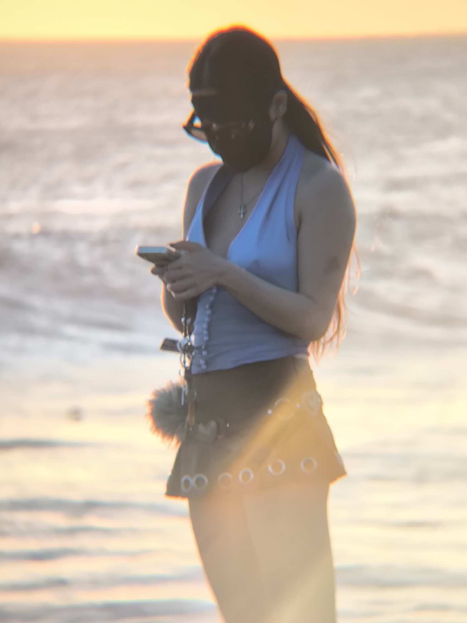 A woman stands on a beach, looking at a mobile phone. She wears a light blue halter top, a brown mini skirt with silver rings, and a black hat. She also wears a necklace with a cross. The background shows a body of water and a sunset. The location is Unknown, Unknown.