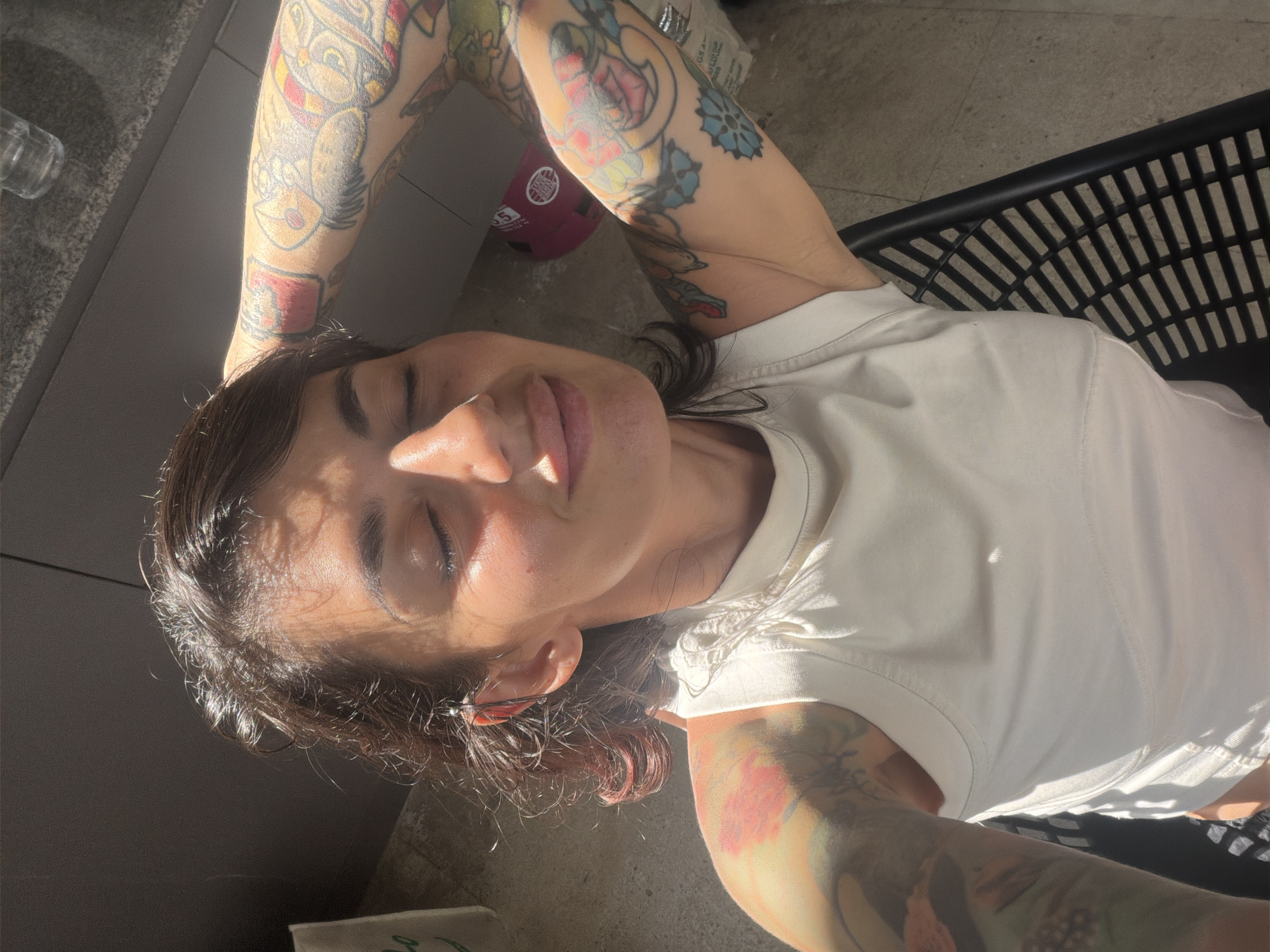 The image depicts an adult female positioned outdoors or in a brightly sunlit interior, in Kuta Selatan, Indonesia. She is seen from an overhead angle, leaning back with her eyes closed and a slight smile. Her dark brown, wavy hair, with visible reddish tones on the viewer's left, is illuminated by direct sunlight. She has extensive, colorful tattoos covering both arms, forming full sleeves. She is wearing an off-white sleeveless tank top. A dark, wire-like earring is visible in her left ear. Both of her arms are raised, with her hands resting behind her head. The background features a light gray, possibly tiled floor, a dark gray vertical surface on the viewer's left, and the slatted black backrest of a chair in the lower right. A clear plastic bottle is visible on a dark surface in the upper right, and a red object lies on the floor in the mid-right.