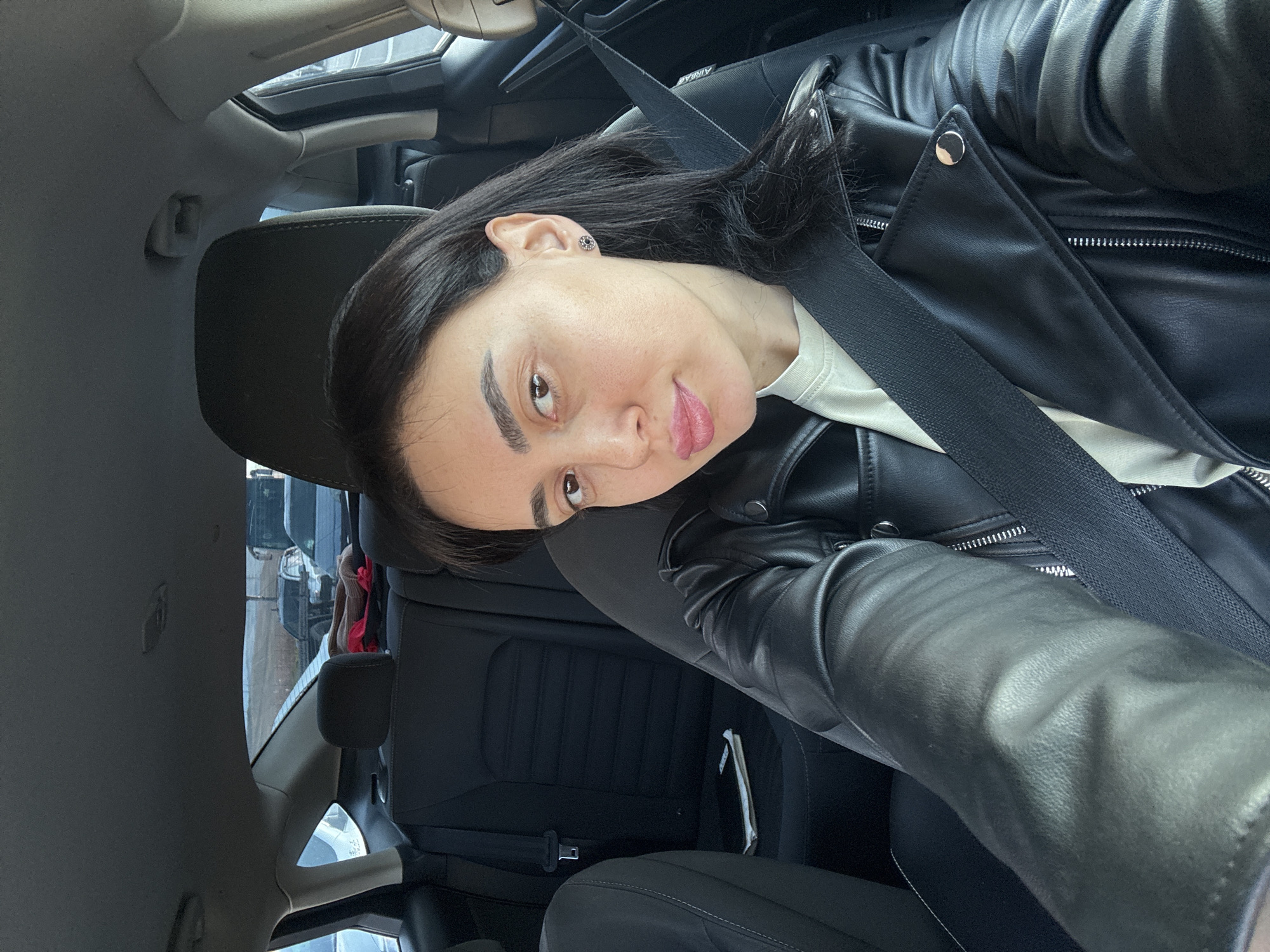 A woman sits in the driver's seat of a car, wearing a black leather jacket and a white shirt, with a seatbelt fastened across her chest. The interior of the car is black and grey. A sunroof is visible above. Outside the car, a blurred urban scene suggests a city environment. Without explicit landmarks or street signs, it is impossible to definitively confirm the location as Tbilisi, Georgia.
