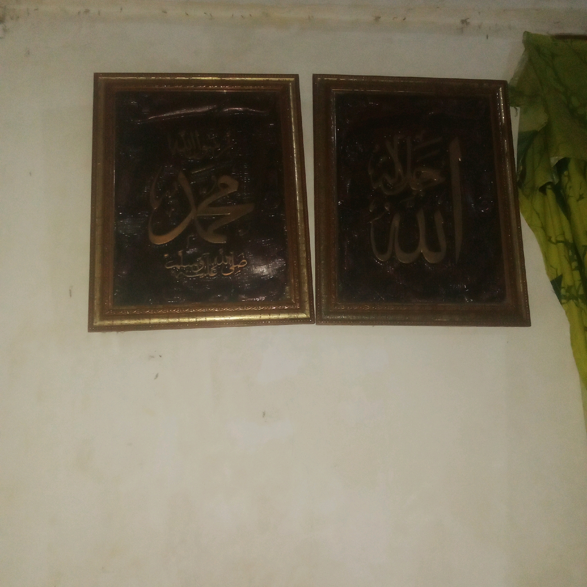 The image shows two framed Islamic calligraphy pieces hanging on a textured, light-colored wall. The left frame contains calligraphy that appears to spell "Muhammad" in Arabic script. Below this, there is smaller Arabic text, possibly a salutation. The right frame contains calligraphy that appears to spell "Allah" in Arabic script. Both frames have a dark background with raised, gold-colored lettering. They are positioned side-by-side, with a slight gap between them. To the right of the frames, a portion of a green fabric with a patterned texture is visible, hanging vertically. The wall itself has some minor imperfections and dust marks. The lighting suggests an indoor setting, possibly a home or a place of worship. Without further information or visible landmarks, the specific location in Karawang, Indonesia, cannot be determined.